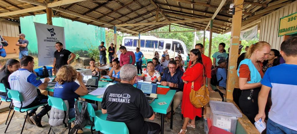 foto colorida mostra as costas de quatro servidores da Vara da Justiça Itinerante sentados em frente a mesas com notebooks realizando atendimentos. A frente está a comunidade aguardando o atendimento, alguns em pé e outros sentados.