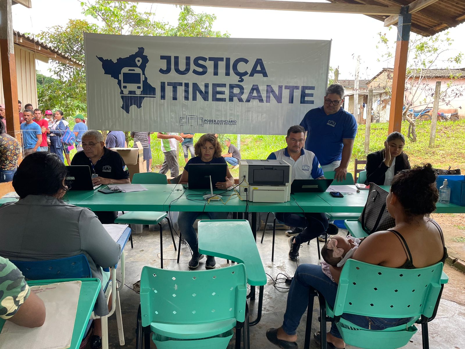 Foto colorida mostra quatro servidores da Vara da Justiça Itinerante sentados em frente a mesas com notebooks realizando atendimentos. Outro servidor da Vara da Justiça Itinerante está em pé auxiliando um dos servidores no atendimento. Frente a eles estão duas mulheres sentadas em cadeiras verdes. Um banner escrito “JUSTIÇA ITINERANTE. PODER JUDICIÁRIO DE RORAIMA” está hasteado entre duas colunas de madeira atrás dos servidores. 