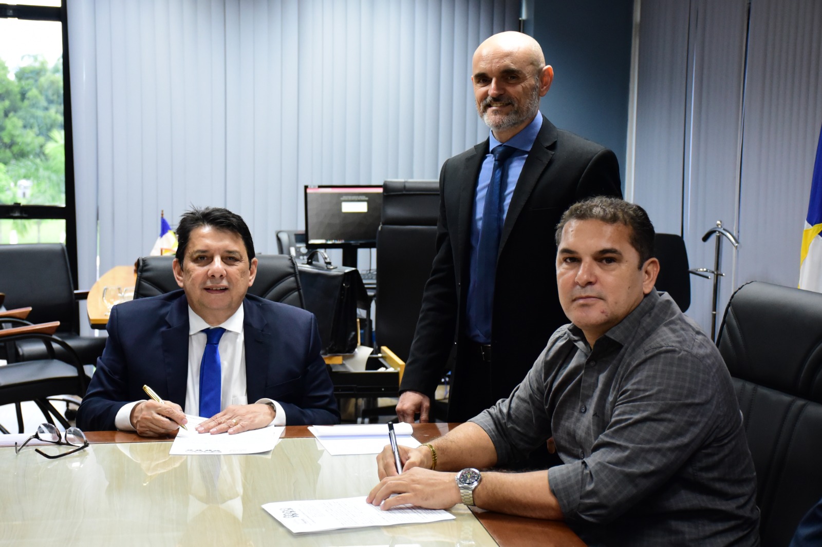 foto colorida mostra o presidente do TJRR, desembargador Jésus Nascimento, na mesa de reunião da sala da presidência do TJRR e  o presidente da Caer, James Serrador, assinando o termo de cooperação técnica entre o Tribunal de Justiça de Roraima e a Caer. Ambos estão sentados na mesa olhando para a câmera, ao lado do presidente, em pé olhando para a câmera está o juiz Air Marin. Atrás deles estão objetos como sofás, mesas, quadros e persianas.
