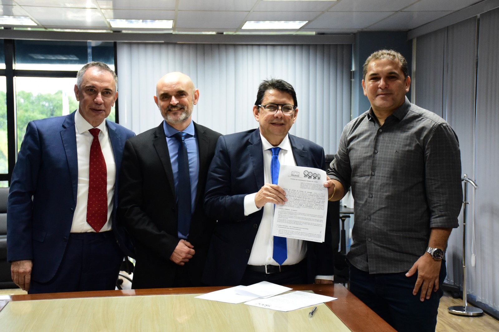 foto colorida mostra o presidente do TJRR, desembargador Jésus Nascimento, e o presidente da Caer, James Serrador, segurando  o termo de cooperação técnica entre o Tribunal de Justiça de Roraima e a Caer. Ao lado do presidente está o juiz Air Marin e o desembargador Almiro Padilha, todos estão em pé, posando para a foto. Atrás deles estão objetos como sofás, mesas, quadros e persianas.