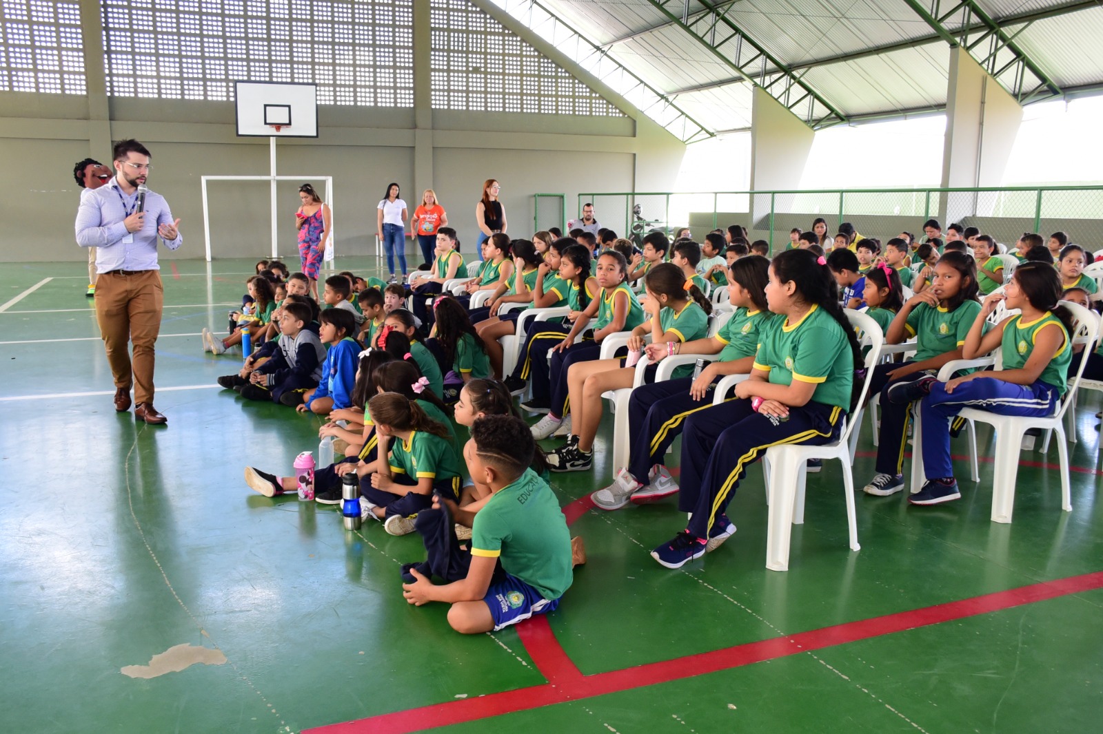 foto colorida mostra a direita um grupo de alunos da Escola Municipal Jael Barradas sentados em cadeiras e no chão do ginásio da instituição acompanhando a palestra que está sendo ministrada pelo servidor do TJRR, Josué os.Ferreira. O palestrante está de pé, ao lado esquerdo da foto, segurando o microfone e gesticulando com uma das mã