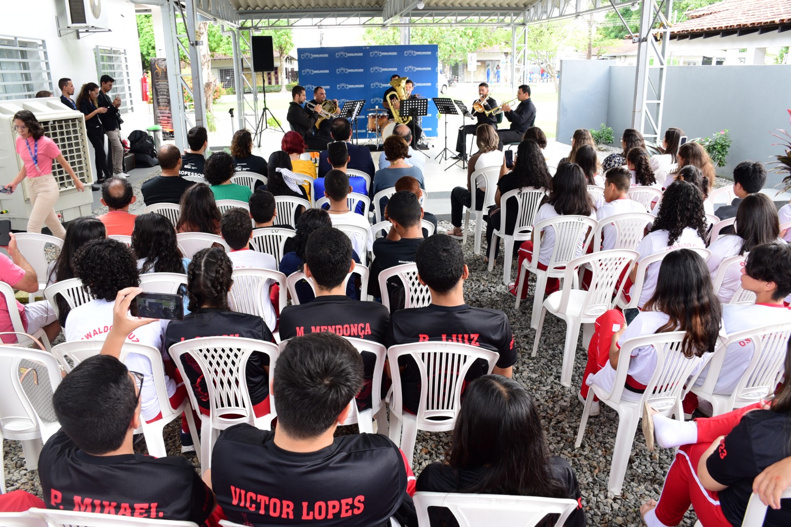 FOTO COLORIDA DE UMA TURMA DE ALUNOS NA AREA EXTERNA DO CMC SENTADOS OBSERVANDO A APRESENTAÇÃO DE MUSICA CLASSICA