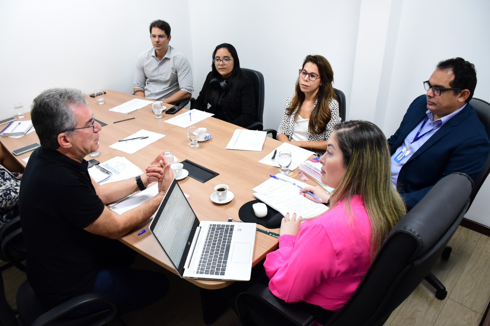 foto colorida mostra a juíza Rafaella Holanda sentada na ponta de uma mesa retangular juntamente a outras cinco pessoas durante reunião. Todos estão sentados ao redor da mesa, que em sua superfície estão objetos como papéis, canetas,copos, xícaras, pastas e um notebook.