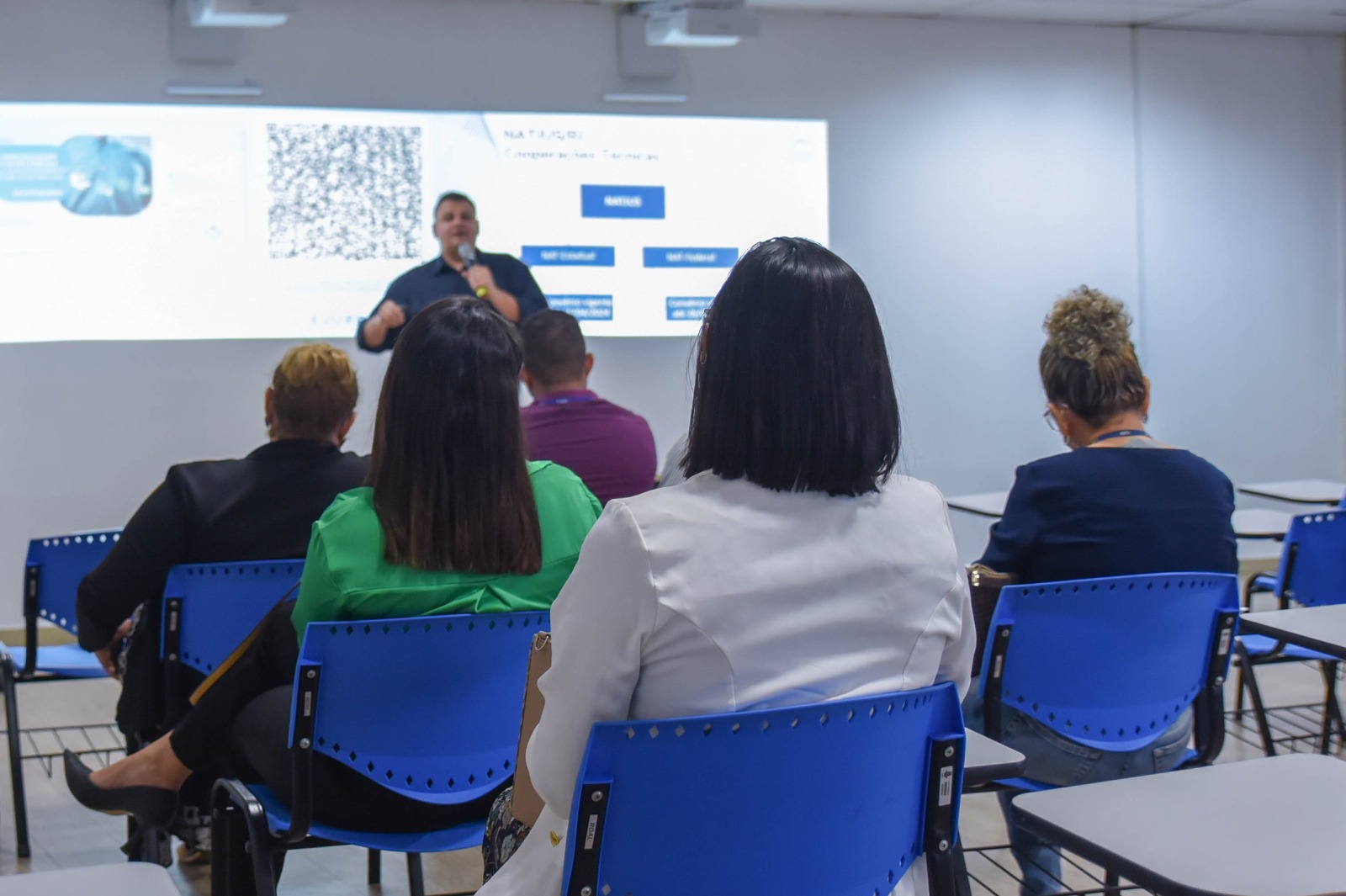FOTO COLORIDA DE UMA SALA DE AULA E A FRENTE UM PROFESSOR FALANDO AO MICROFONE