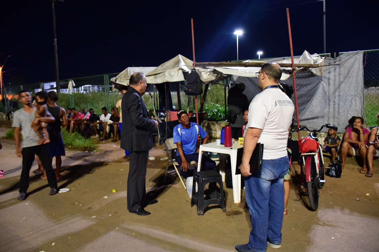  foto colorida mostra servidores do TJRR e TRT em pé, à noite, falando com dois migrantes que estão sentados ao redor de uma mesa de plástico. Eles estão nas proximidades da rodoviária de Boa Vista.
