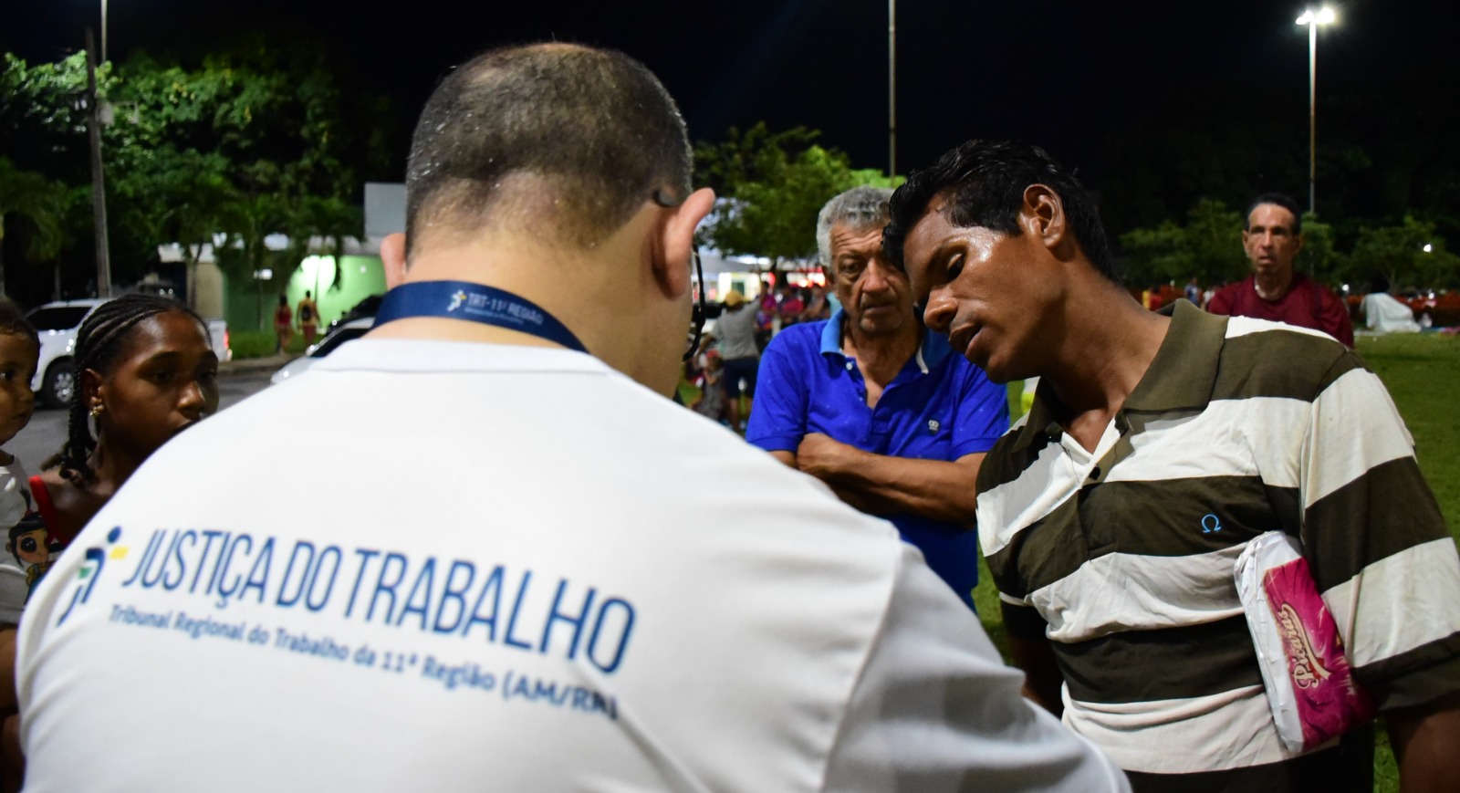 foto colorida mostra o servidor do TRT em pé, de costas, falando com um grupo de três migrantes em frente a rodoviária de Boa Vista.