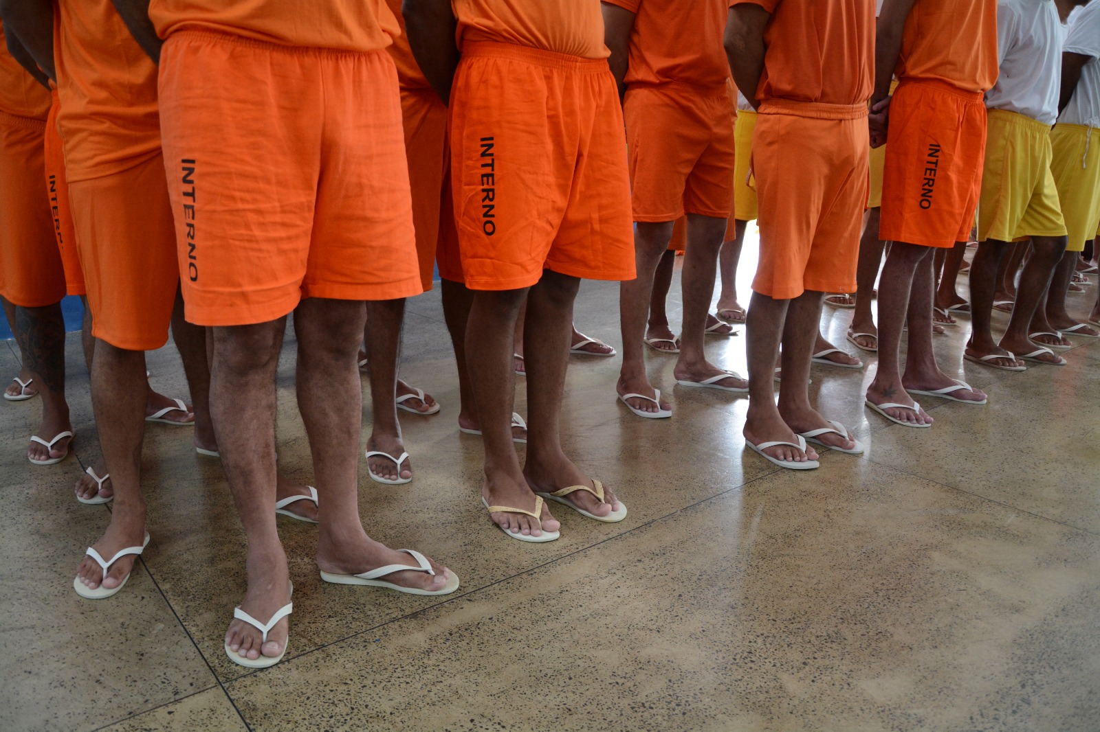 foto colorida dando enfoque da cintura para baixo de homens enfileirados em pé, com vestimentas laranjas e de chinelos branco. Abaixo está o texto “SISTEMA PENITENCIÁRIO Tribunal de Justiça de Roraima participa de mutirão carcerário que visa desafogar unidades prisionais em todo país”