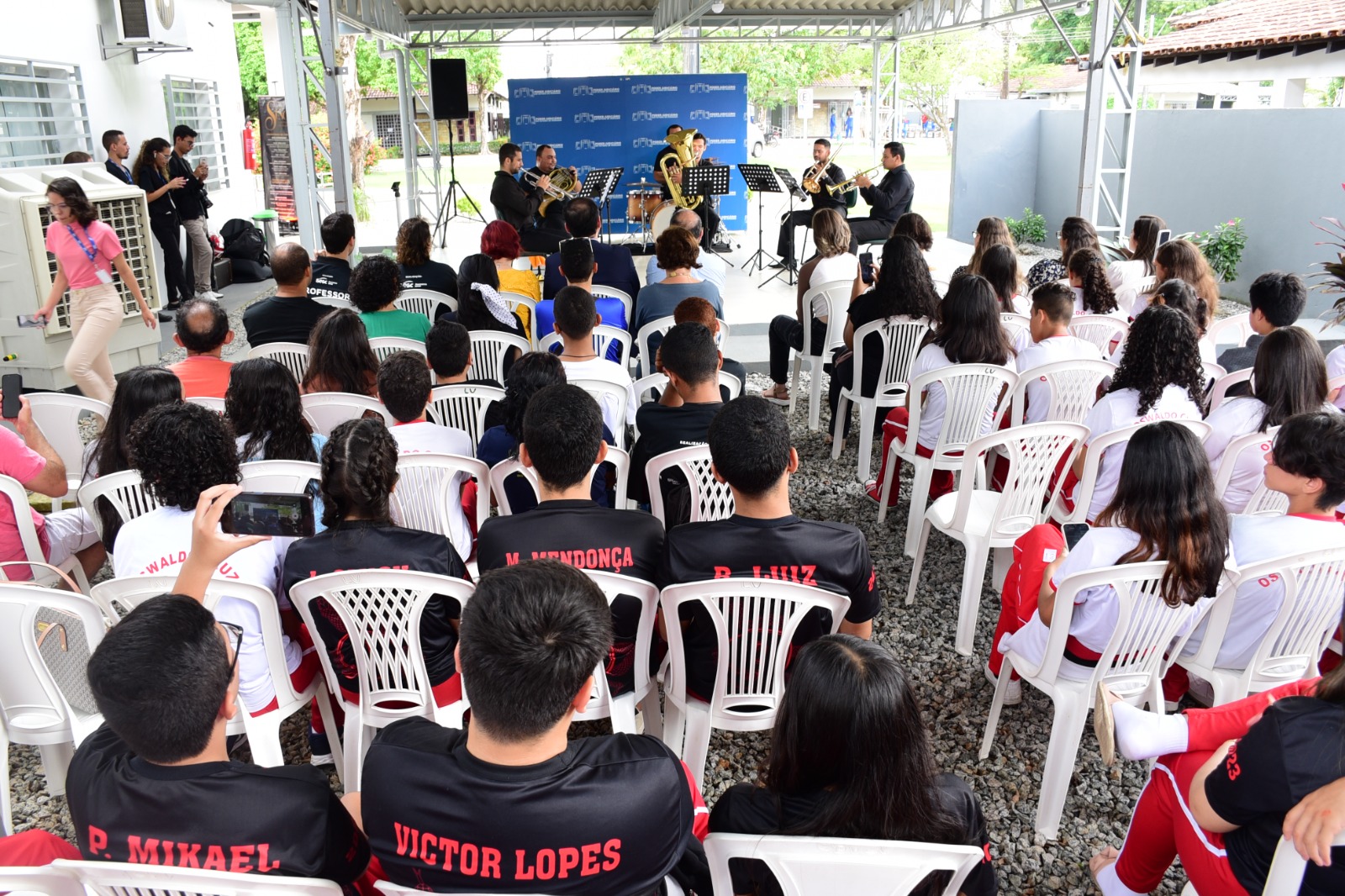 foto colorida da area externa do cmc. Nela há alunos do colégio sentados em cadeira de plástico prestando atenção na orquestra que estava apresentando