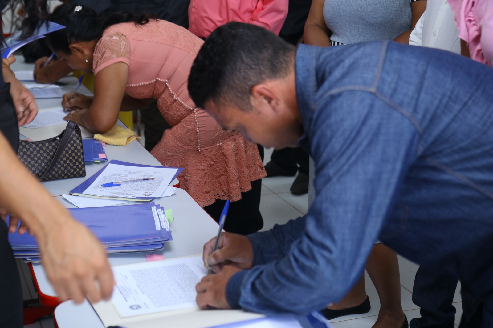  foto colorida mostra homem assinando documento em cima de uma mesa, durante a cerimônia do casamento coletivo da Justiça itinerante, do TJRR. Logo mais no fundo da imagem, mostra uma mulher assinando documento em cima da mesma mesa.