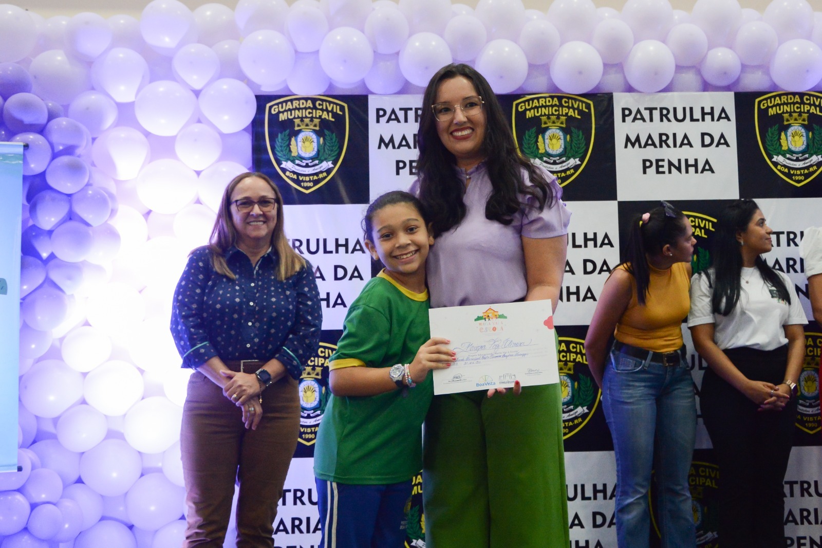 oto colorida com a Juíza titular da Coordenadoria Estadual da Mulher em Situação de Violência Doméstica e Familiar do TJRR, Suellen Alves, entregando o certificado para aula no ensino fundamental. Ao fundo tem balões roxos junto com backdrop que possui emblema da Guarda Municipal Civil e escrito “PATRULHA MARIA DA PENHA”