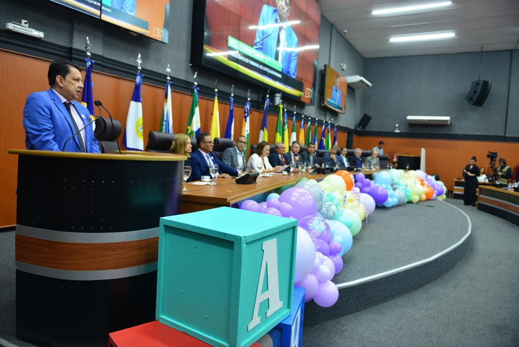 foto colorida mostrando o Presidente do TJRR, desembargador Jésus Nascimento no pulpito, ao lado está a mesa de autoridades compostas por representantes do Legislativo, Executivo e Judiciário. A frente da mesa contém balões roxos, azul, amarelo e laranja em tons claros.