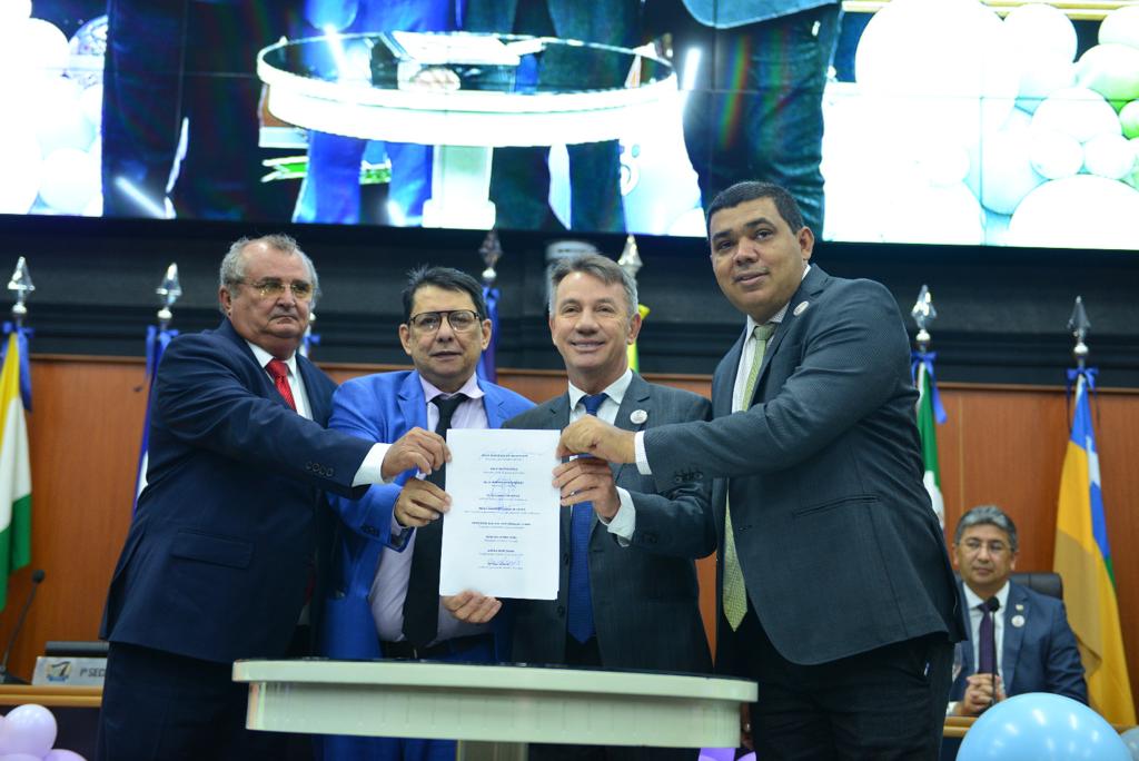 foto colorida mostrando o Presidente do TJRR, desembargador Jésus Nascimento no pulpito, ao lado está a mesa de autoridades compostas por representantes do Legislativo, Executivo e Judiciário. A frente da mesa contém balões roxos, azul, amarelo e laranja em tons claros.