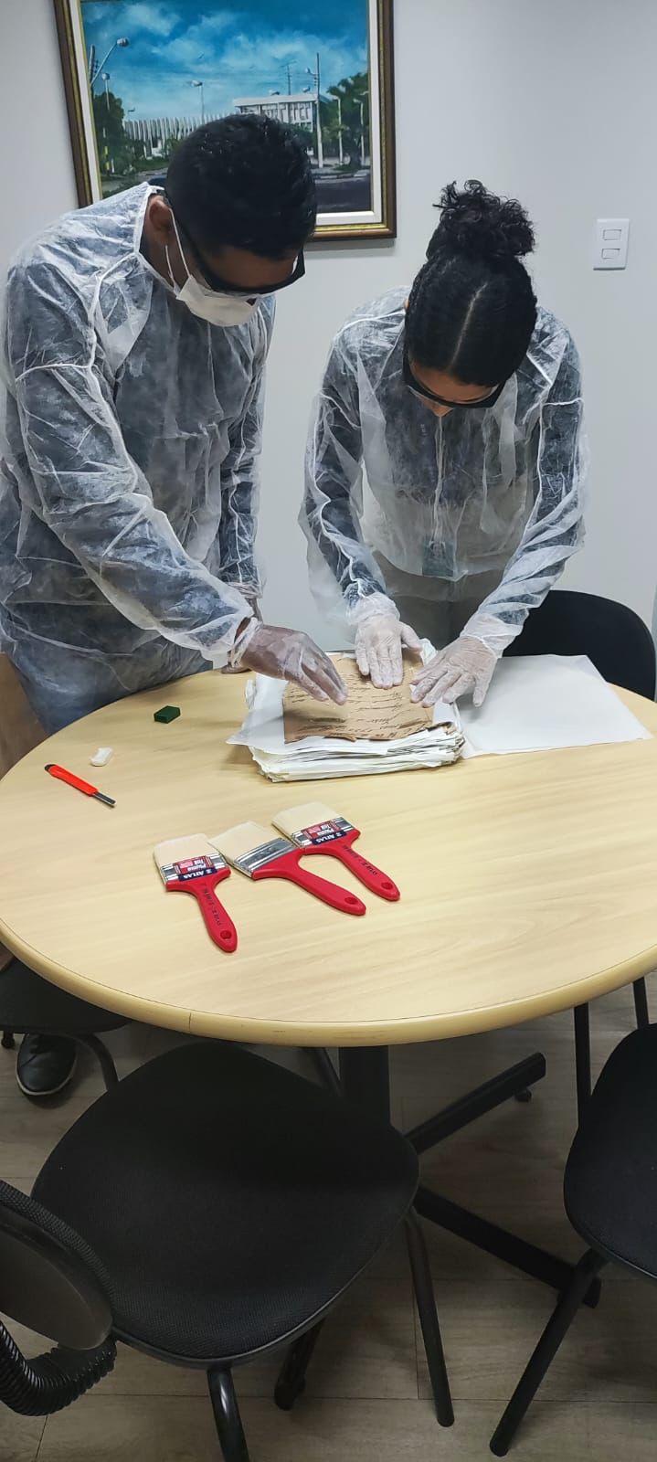 oto colorida na vertical mostrando duas pessoas da equipe do Centro de Memória e Cultura, realizando o processo de higienização nos documentos sob uma mesa redonda com pincéis grandes e vermelhos.  Ambos estão  de máscaras brancas, óculos e jaleco de proteção e luva branca