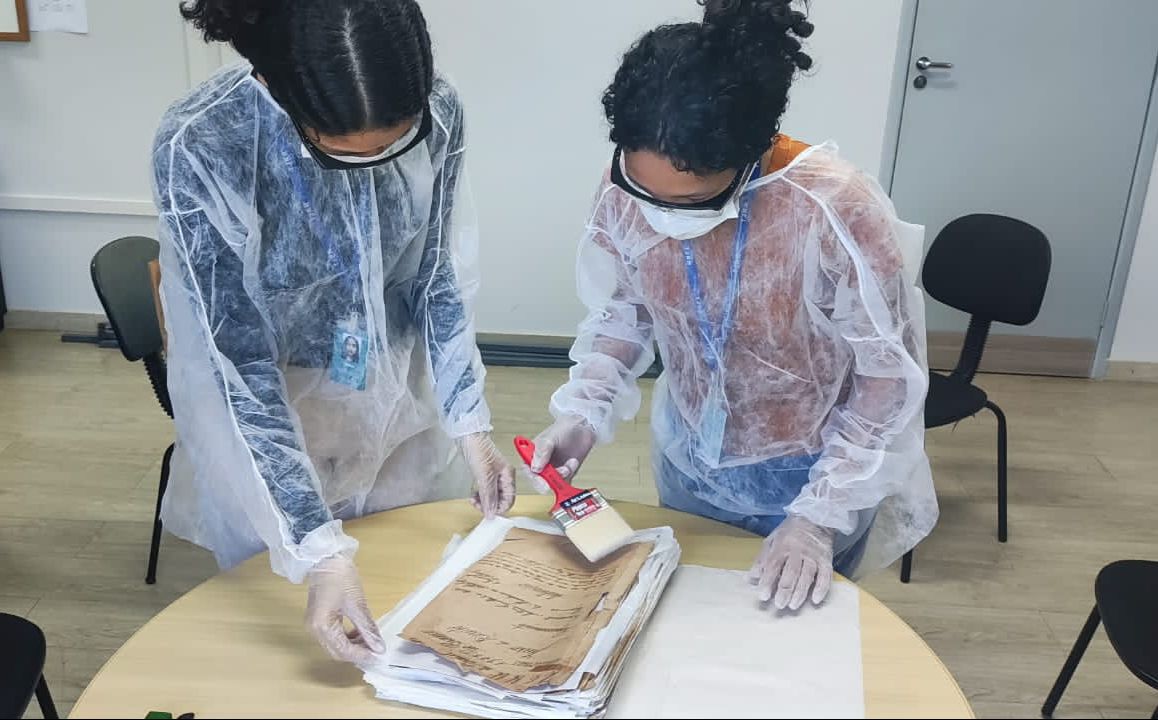 foto colorida mostrando duas pessoas da equipe do Centro de Memória e Cultura, realizando o processo de higienização nos documentos sob uma mesa redonda com pincéis grandes e vermelhos.  Ambos estão  de máscaras brancas, óculos e jaleco de proteção e luva branca