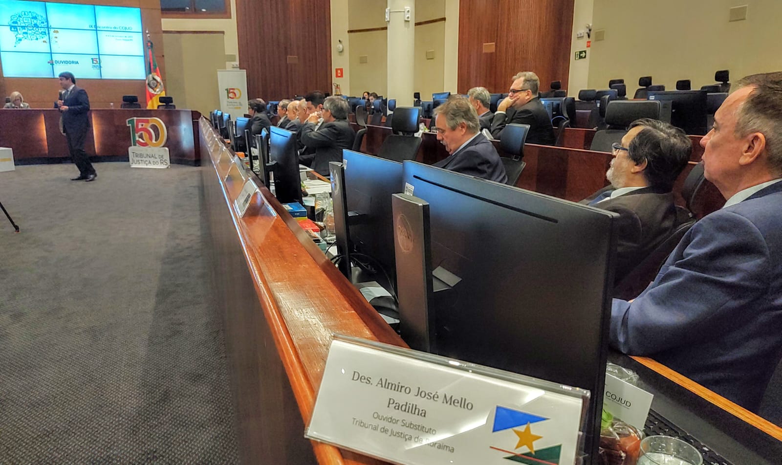 A imagem colorida contém os ouvidores judiciais do país, sentados reunidos durante o IX Encontro do Colégio Nacional de Ouvidores Judiciais - COJUD, enquanto um ouvidor está palestrando. Abaixo a frase “EVENTO NACIONAL - Desembargador Almiro Padilha representa Ouvidoria do TJRR no Encontro de Ouvidores.”