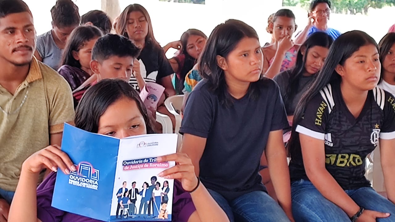 foto colorida dando enfoque em uma adolescente indigena lendo a cartilha da ouvidoria enquanto ao fundo é possivel ver o restante da comunidade presente