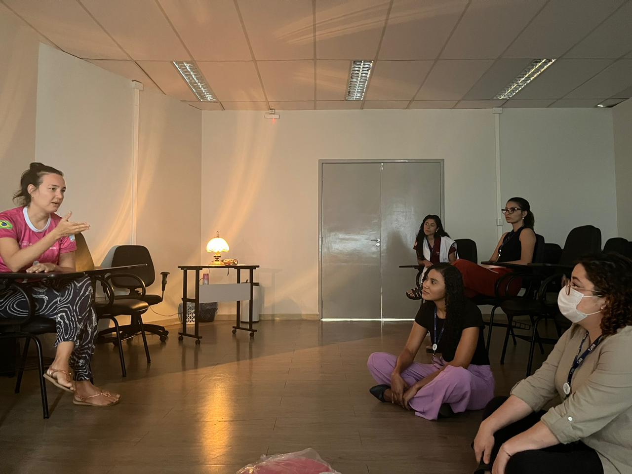 Imagem colorida horizontal de uma sala clara parcialmente iluminada por um abajur de luz amarela. Ao todo, na sala, estão 5 mulheres, uma sentada em uma cadeira do lado esquerdo da foto, duas à sua frente sentadas no chão e outras duas sentadas atrás dessas em outras cadeiras.