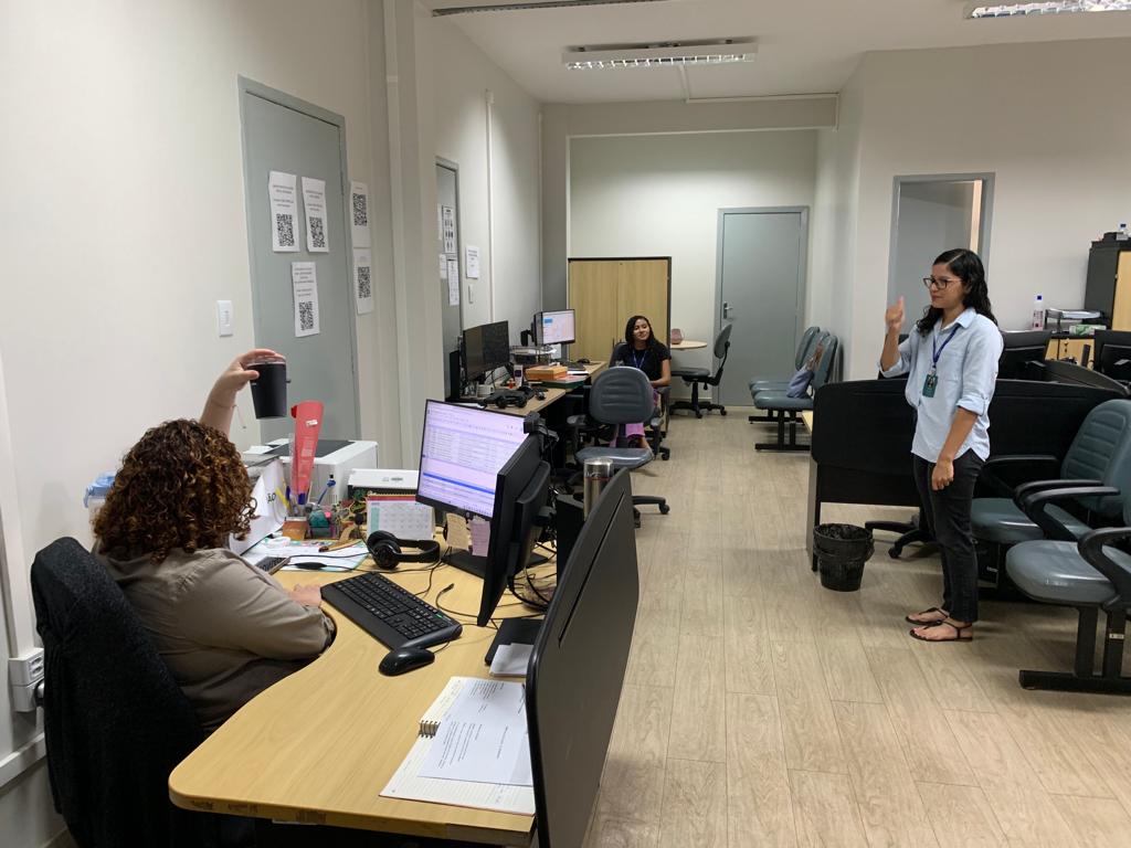 Foto colorida de três mulheres em uma sala. Uma está em pé, e outras duas sentadas nas suas mesas, com os computadores ligados.