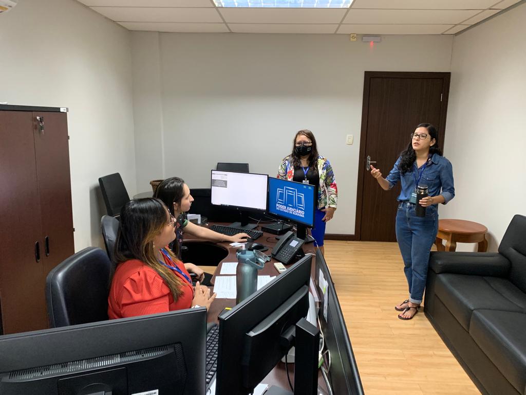  Foto colorida mostra duas mulheres em pé em uma sala, um homem sentado de costas, de frente para alguns monitores, é uma mulher sentada ao fundo.