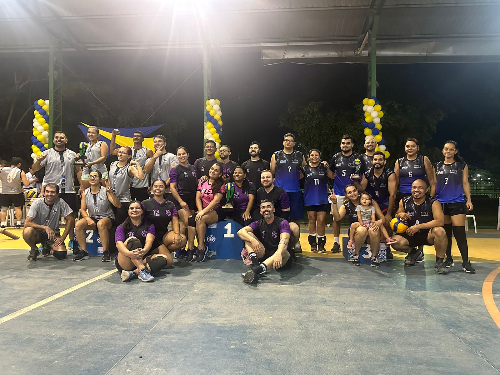 Imagem colorida de atletas com diferentes uniformes reunidos em uma quadra. Ao centro, está o Secretário Geral Henrique Tavares, e um banner do 3° VôleiJus.