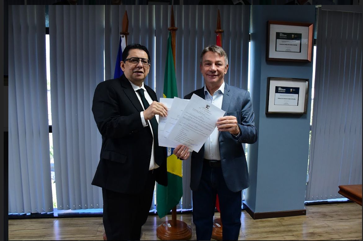foto colorida do presidente do TJRR, desembargador Jésus Nascimento junto com o Governador Antônio denarium pousando para a fotografia om papeis em mãos