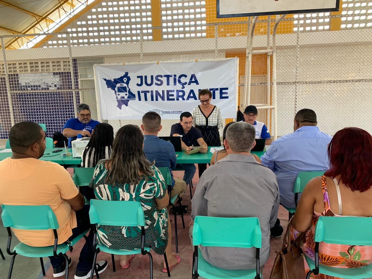 foto colorida na horizontal com oito pessoas sentadas em cadeiras verdes esperando o atendimento. A frente há uma mesa de três servidores realizando o atendimento e atrás há uma banner escrito Justiça Itinerante.