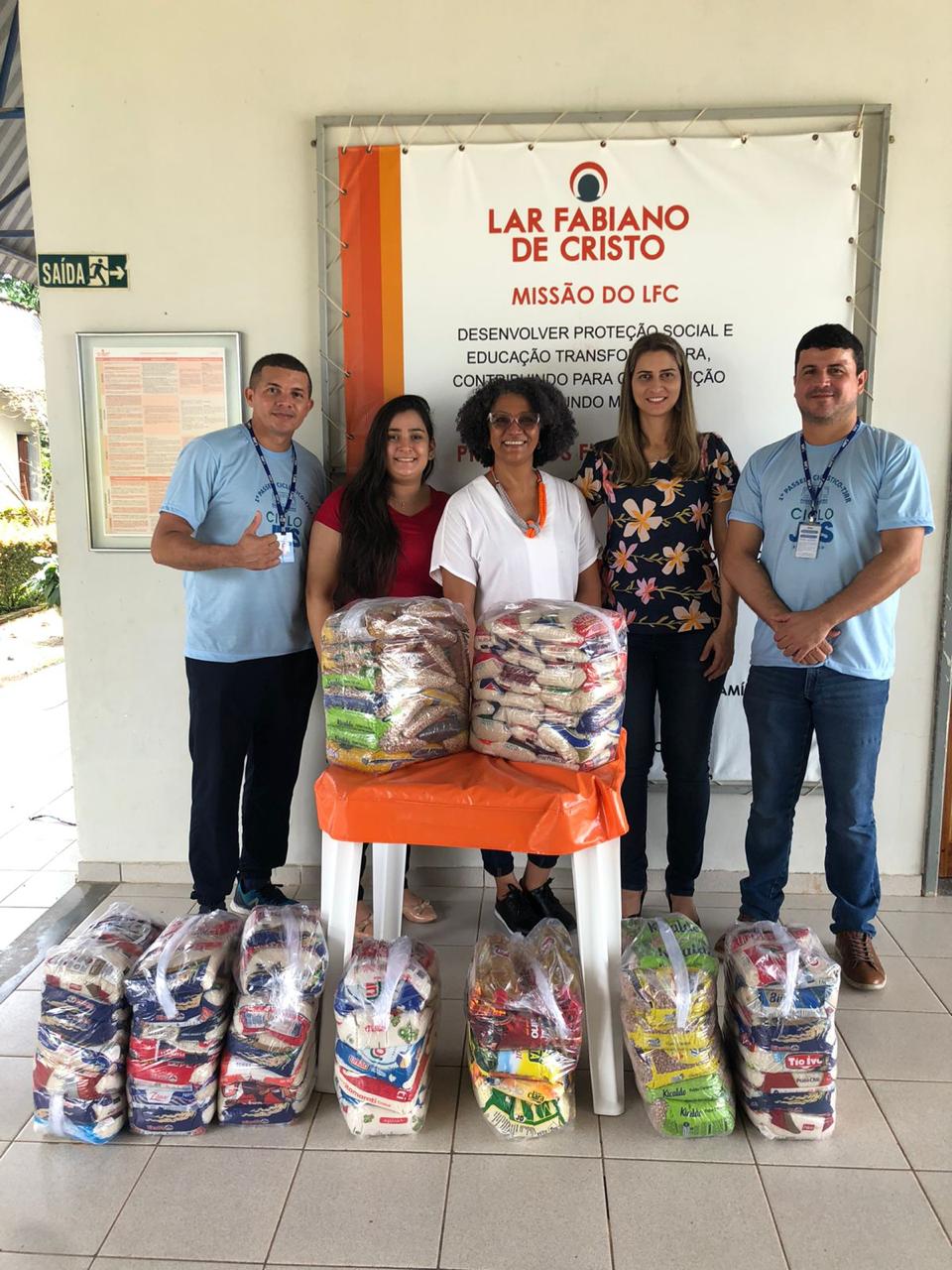 Foto colorida mostrando cinco pessoas em pé posando para foto. A frente deles há sacos cheios de alimentos não perecíveis doados