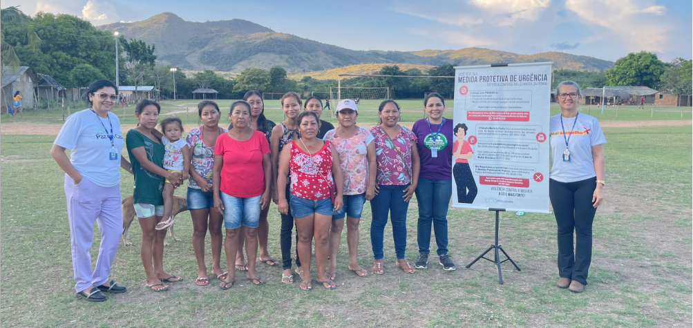 Foto colorida mostra servidores em indígenas de comunidades posando para foto em território indígena, a direita mostrar banner do Programa Maria Vai à Escola.