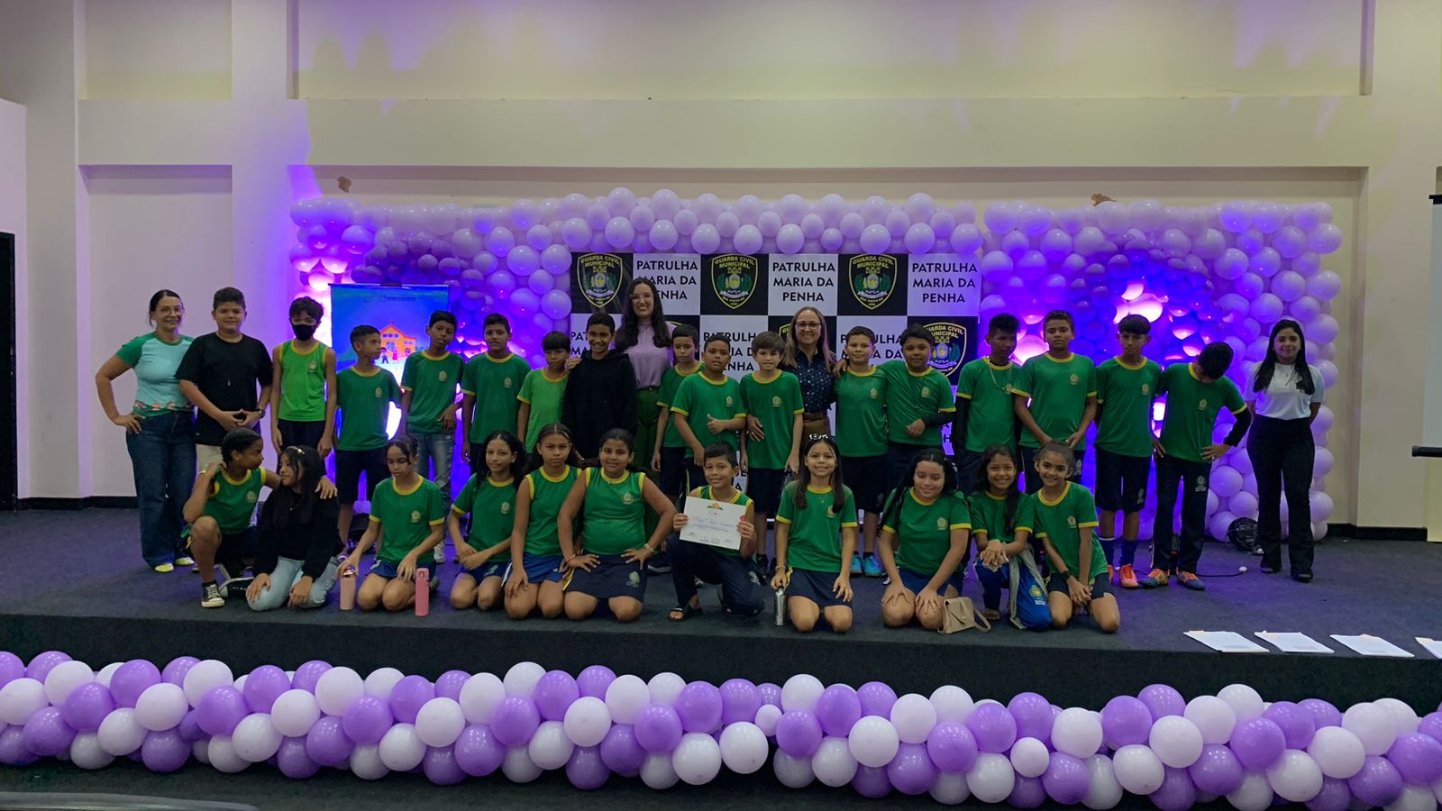 Foto colorida mostra alunos de uma escola posando para foto com certificado em cima de um palco.
