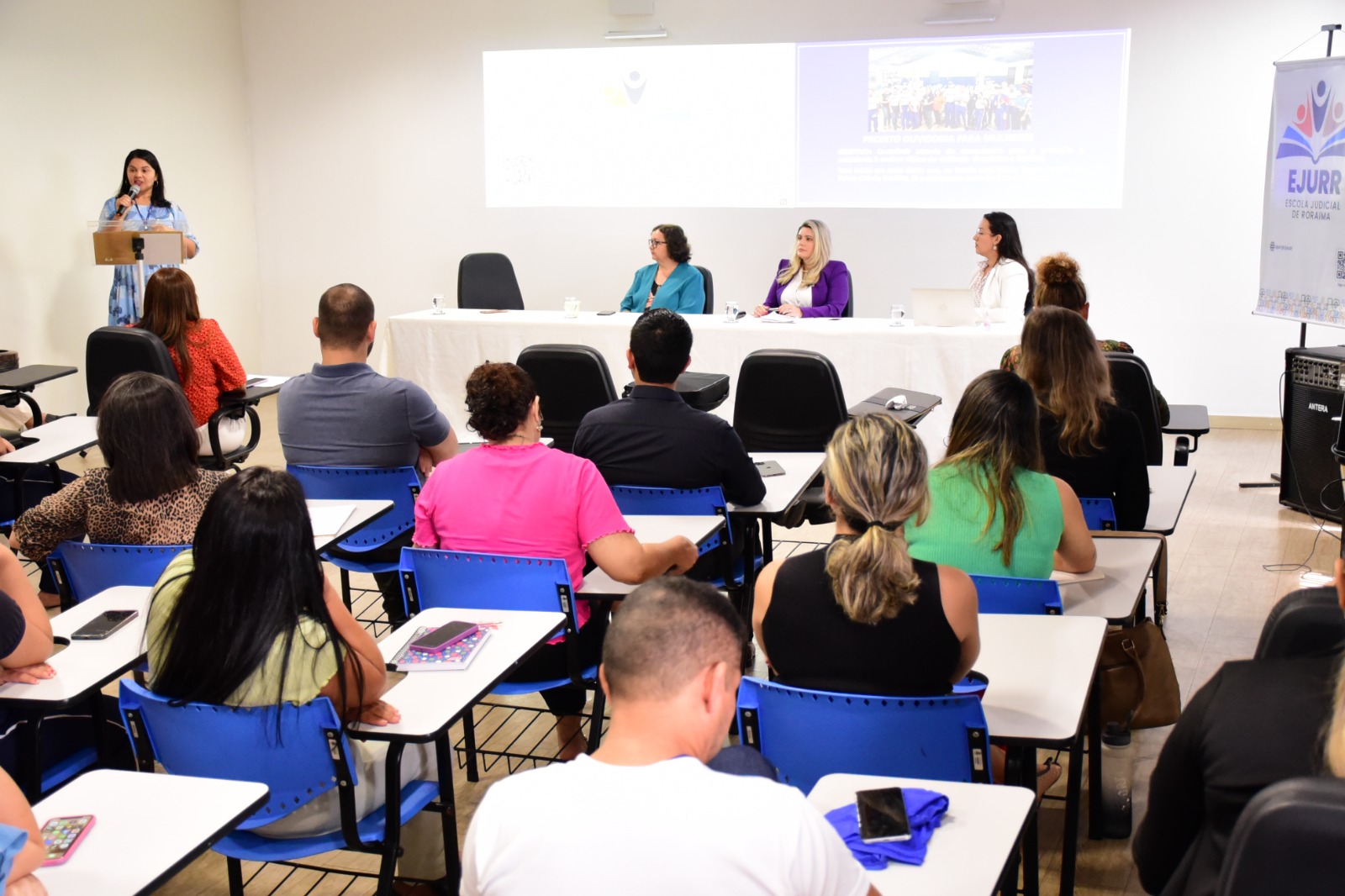 Foto colorida mostra 4 pessoas discursando para alunos em uma sala