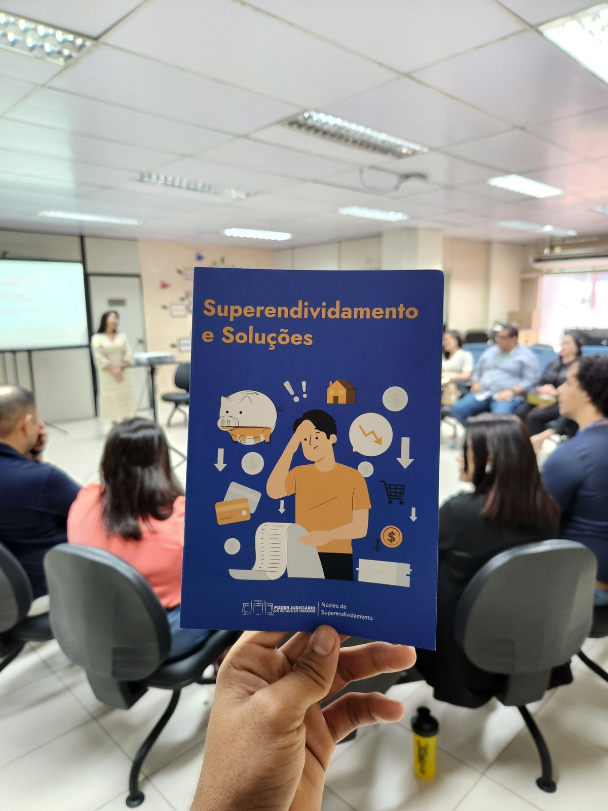 Foto colorida mostra mão segurando um panfleto do curso com a escrita: "Superendividamento e Soluções" durante o curso.
