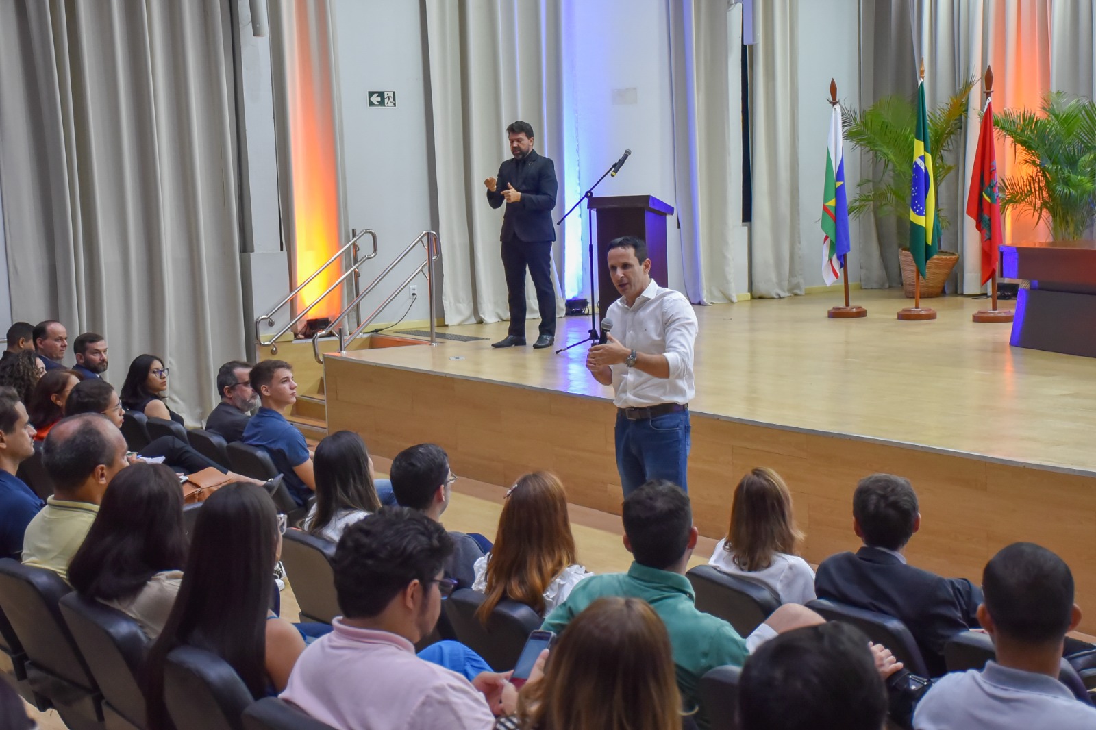 Foto colorida de uma plateia em um auditório assistindo um homem que está falando em pé.