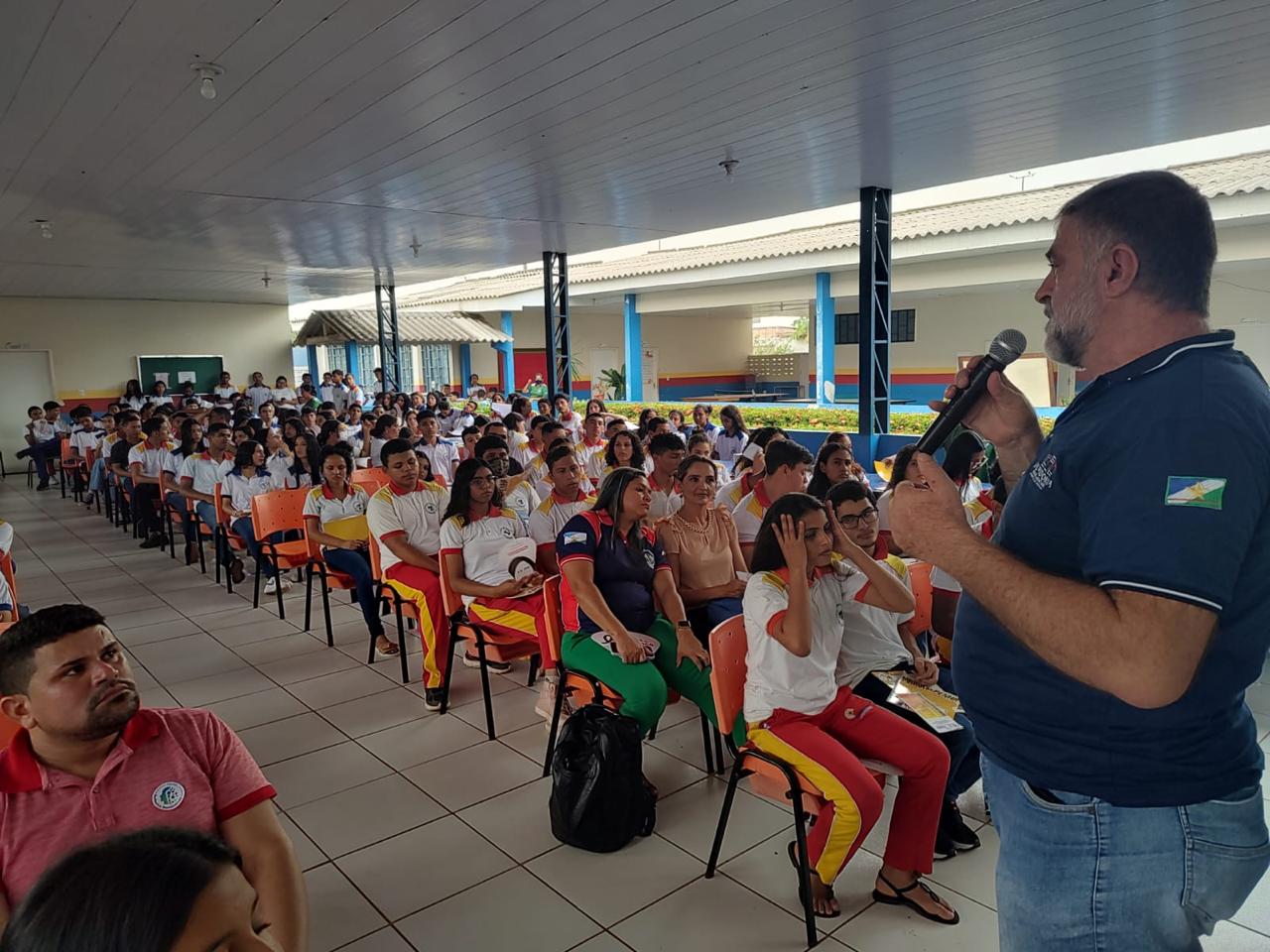 Foto colorida de vários estudantes sentados em cadeiras olhando para o Assistente Técnico da Ouvidoria, Amadeu Triani, que está em pé falando