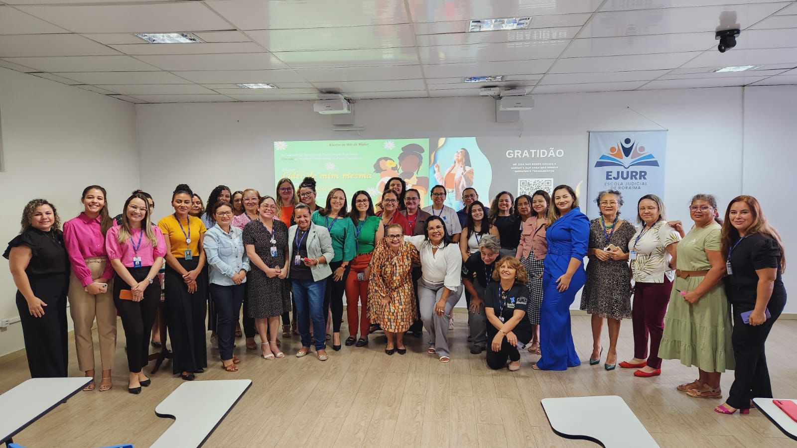 Foto colorida de mulheres uma sala de aula, posando para a foto . Ao fundo, uma palestrante e um intérprete de libras.