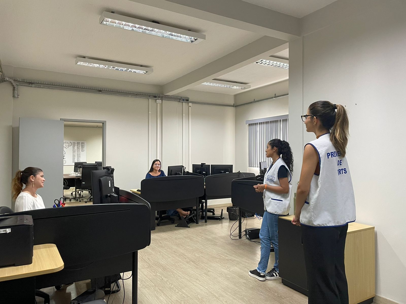 Imagem colorida mostrando pessoas em uma sala. Algumas estão sentadas a mesa e outras em pé.