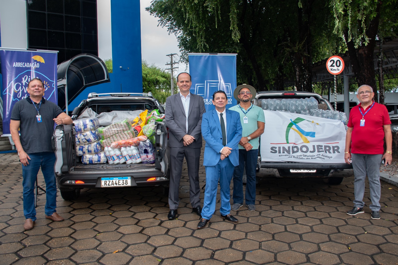 Foto colorida mostrando o o presidente do Tribunal de Justiça de Roraima, desembargador Jésus Nascimento, ao lado do desembargador Cristovão Suter e 3 servidores do TJRR posando para foto. No meio um carro com o porta malas aberto e diversos produtos alimentícios