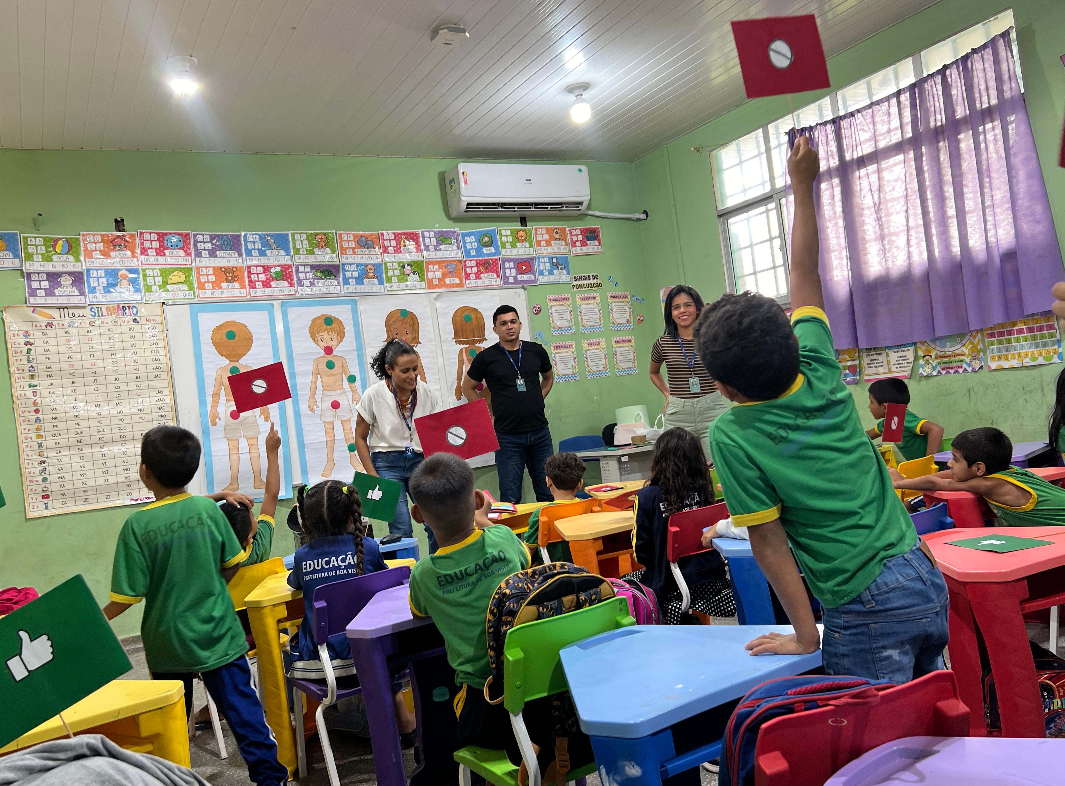 Imagem colorida de uma sala de aula mostrando crianças levantando uma plaquinha durante atividade realizada pela Coordenadoria da Infância e Juventude