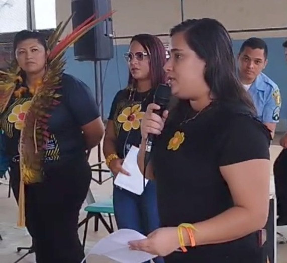 Foto colorida mostrando uma mulher segurando um microfone durante discurso. Atrás dela outras mulheres estão assistindo em pé o discurso