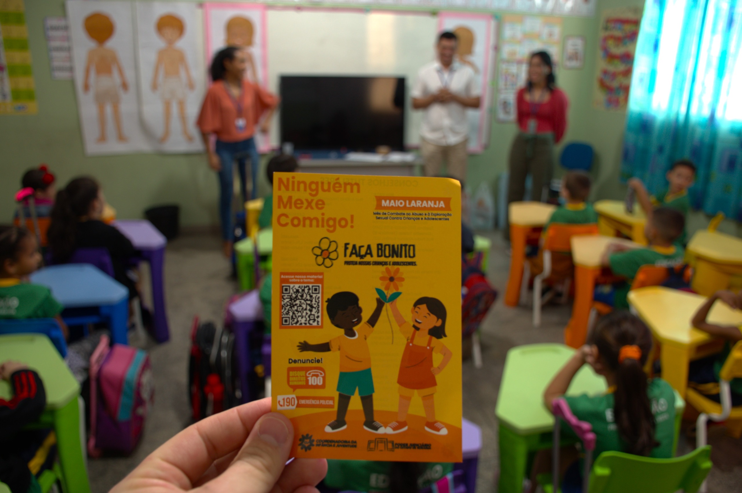 Imagem colorida mostranod uma sala de aula no fundo. Em primeiro plano na imagem um folder da Coordenadoria da Infância e Juventude mostrando duas crianças segurando uma flor. Na imagem o texto "Ninguém mexe comigo!", "Faça Bonito",