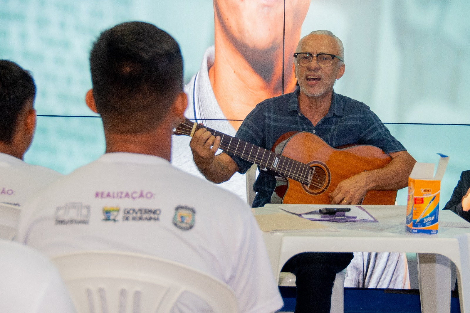 A imagem colorida mostra um artista e cantor local tocando violão e cantando para os visitantes do festival. Abaixo a frase: PRESERVANDO A CULTURA - mãos que escrevem a nossa história é tema do 2° festival de memória e cultura de Roraima