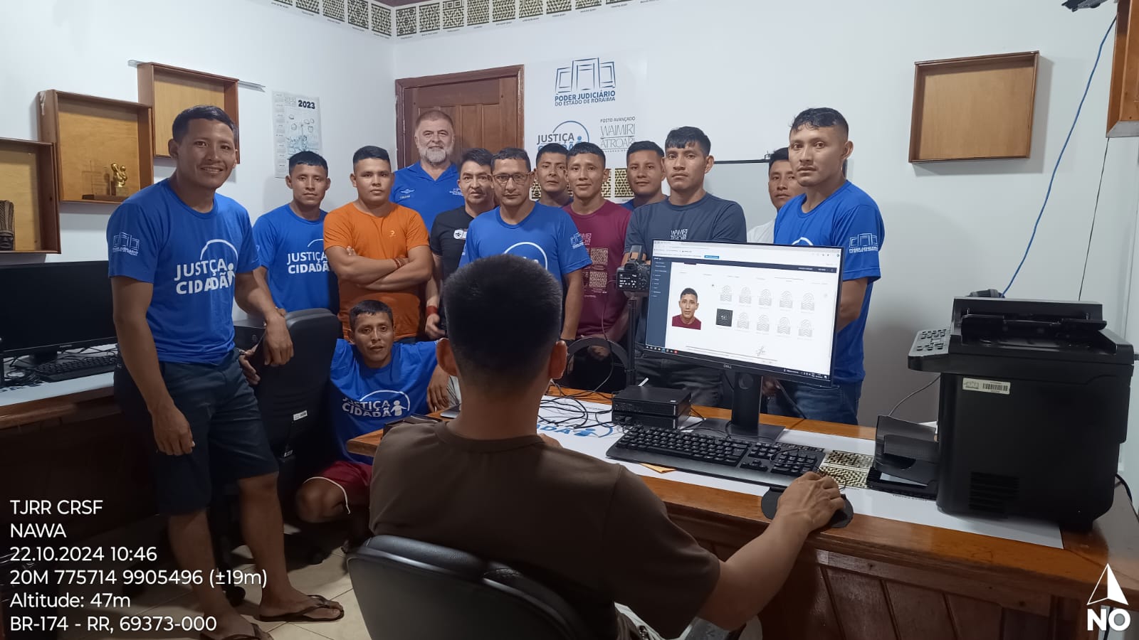 Foto colorida mostra 13 pessoas posando para foto e uma pessoa ao computador trabalhando