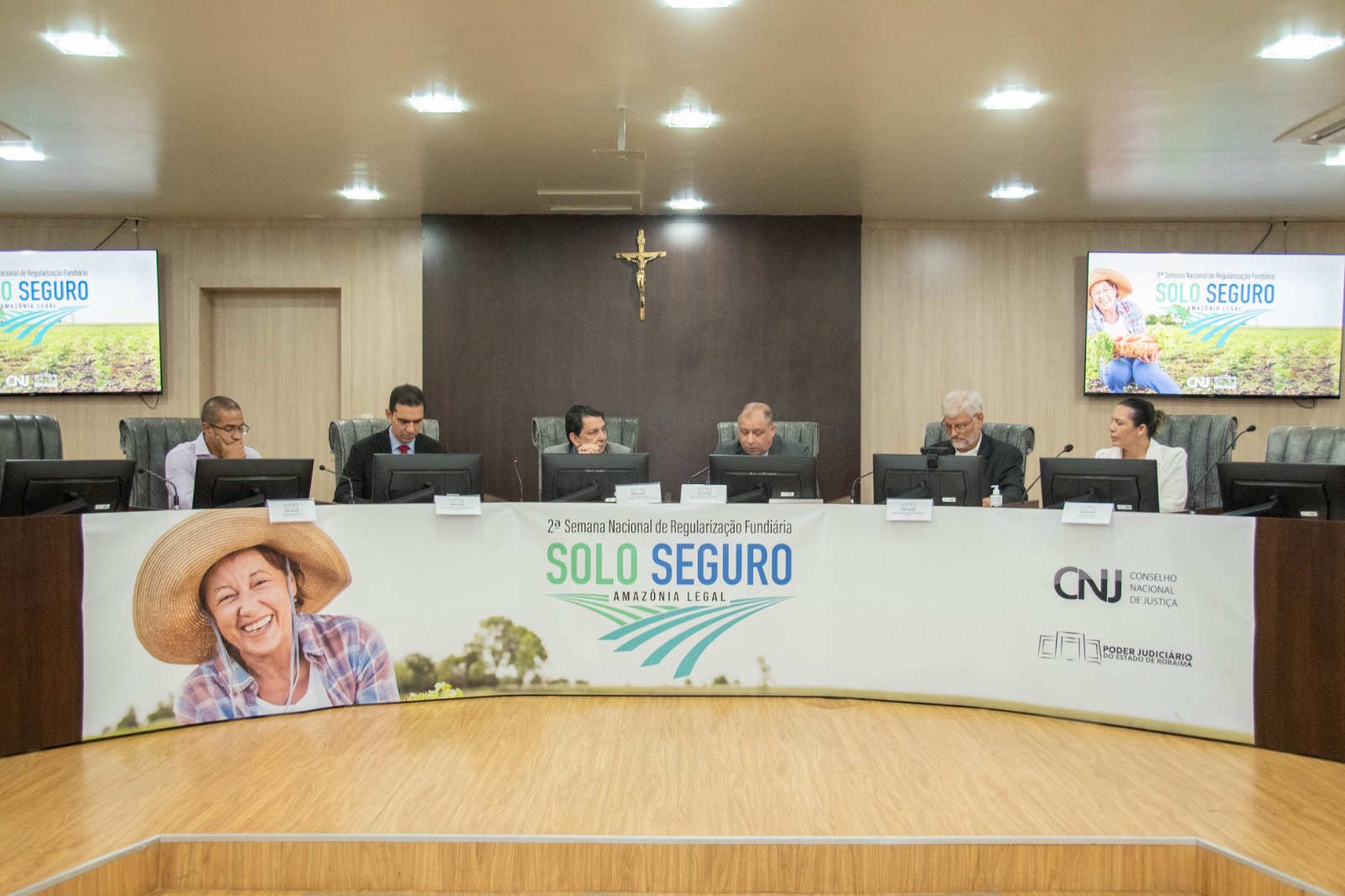 colorida mostrando uma mesa de autoridades, nela se encontram o presidente do TJRR, desembargador Jésus Nascimento, o corregedor-geral de Justiça, desembargador Mozarildo Cavalcanti, e o bispo da Diocese de Roraima, Dom Evaristo Pascoal Spengler. Abaixo na imagem o texto: "SOLO SEGURO - Regularização Fundiária avança em Roraima com doação de terras e entrega de títulos" .