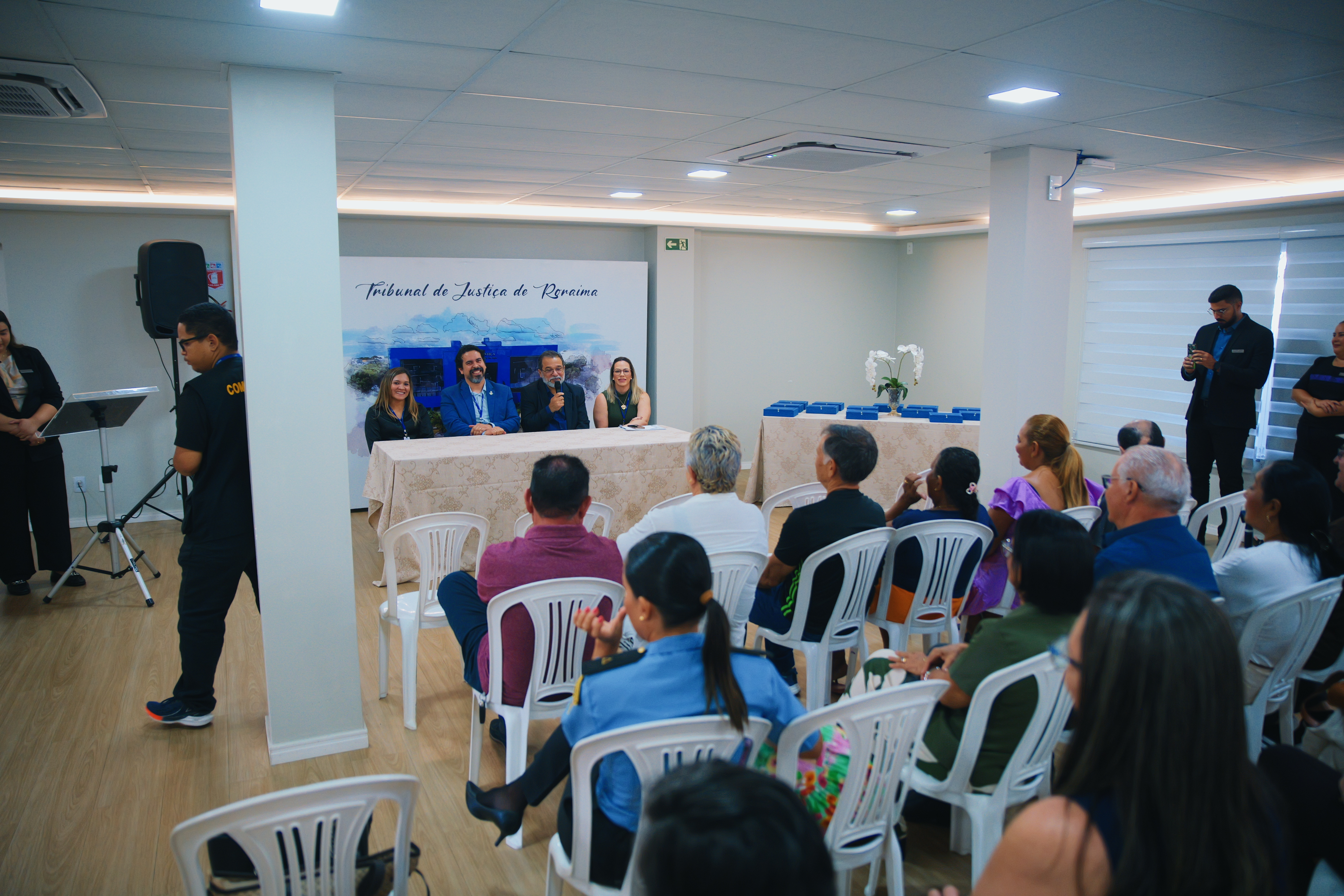 imagem colorida mostra diversas pessoas sentadas de frente para mesa com quatro autoridades do TJRR durante evento em alusão ao dia do aposentado.