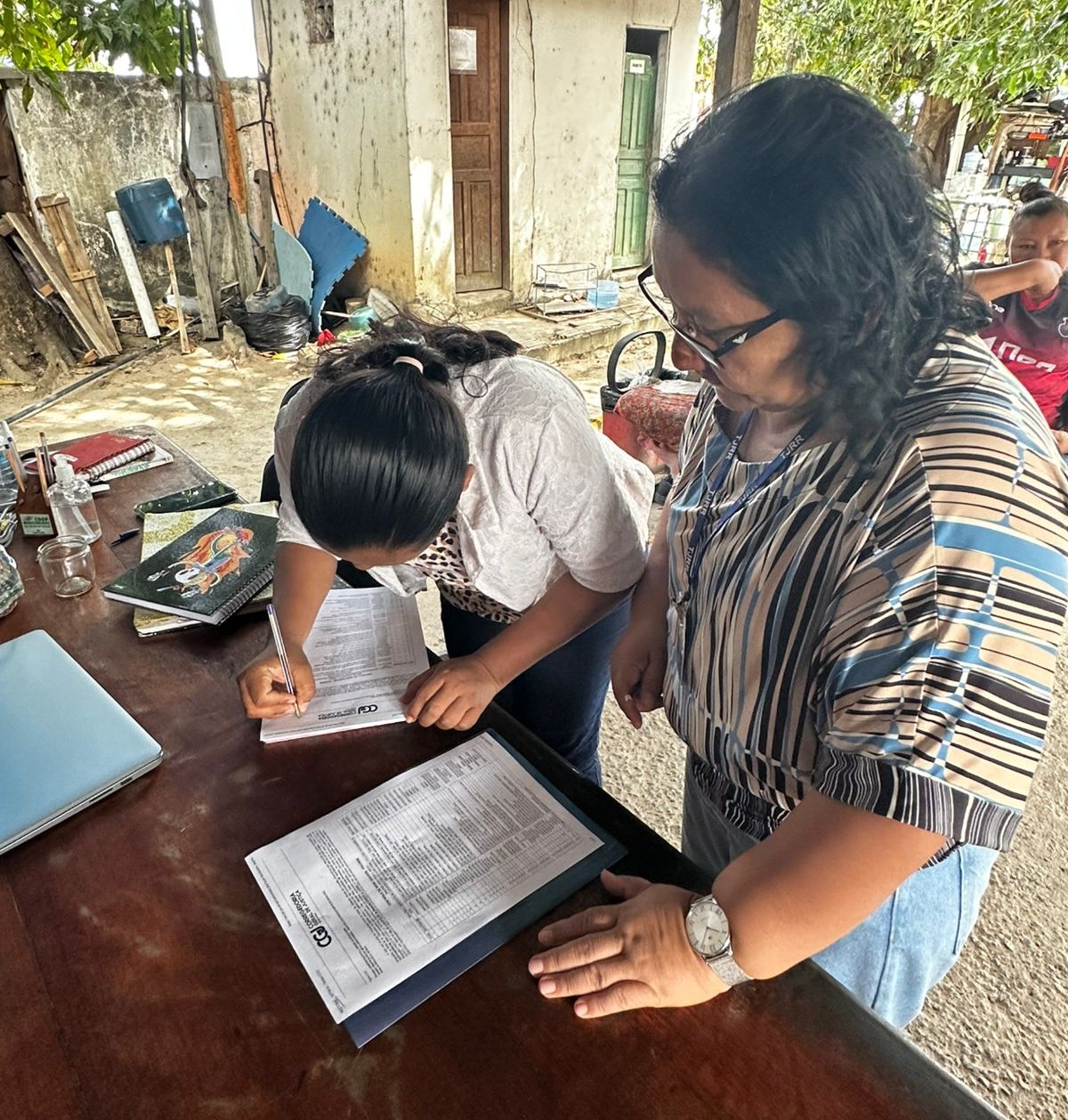 Foto colorida mostra uma mulher inclinada assina um documento sobre uma mesa de madeira. Ao lado, outra mulher de óculos e crachá acompanha a assinatura, com uma pasta e folhas impressas apoiadas na mesa. Há um notebook fechado, celular e cadernos próximos. Ao fundo, parede e portas de uma construção simples e árvores.