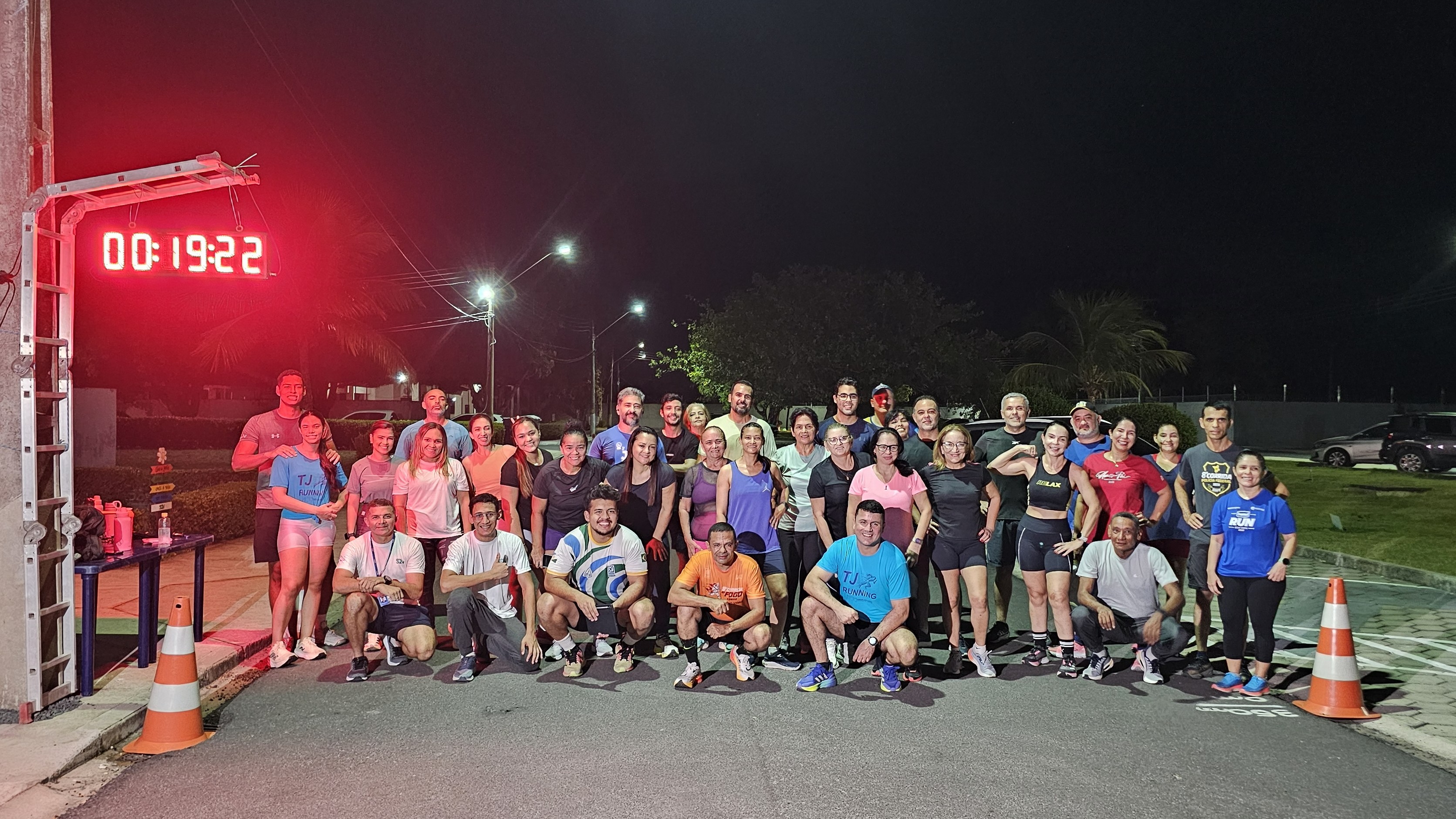 Foto coletiva ao ar livre, à noite, em uma rua iluminada por postes. Um grupo grande de participantes, em roupas de treino, posa em duas fileiras: parte em pé ao fundo e parte agachada/ajoelhada na frente. À esquerda, há um relógio digital vermelho marcando 00:19:22, preso a uma estrutura; abaixo, uma mesa com itens como garrafas e objetos de apoio. No chão, aparecem cones laranja delimitando o espaço. Ao fundo, vê-se gramado, árvores e carros estacionados, sugerindo um ponto de encontro para atividade física do TJRR.