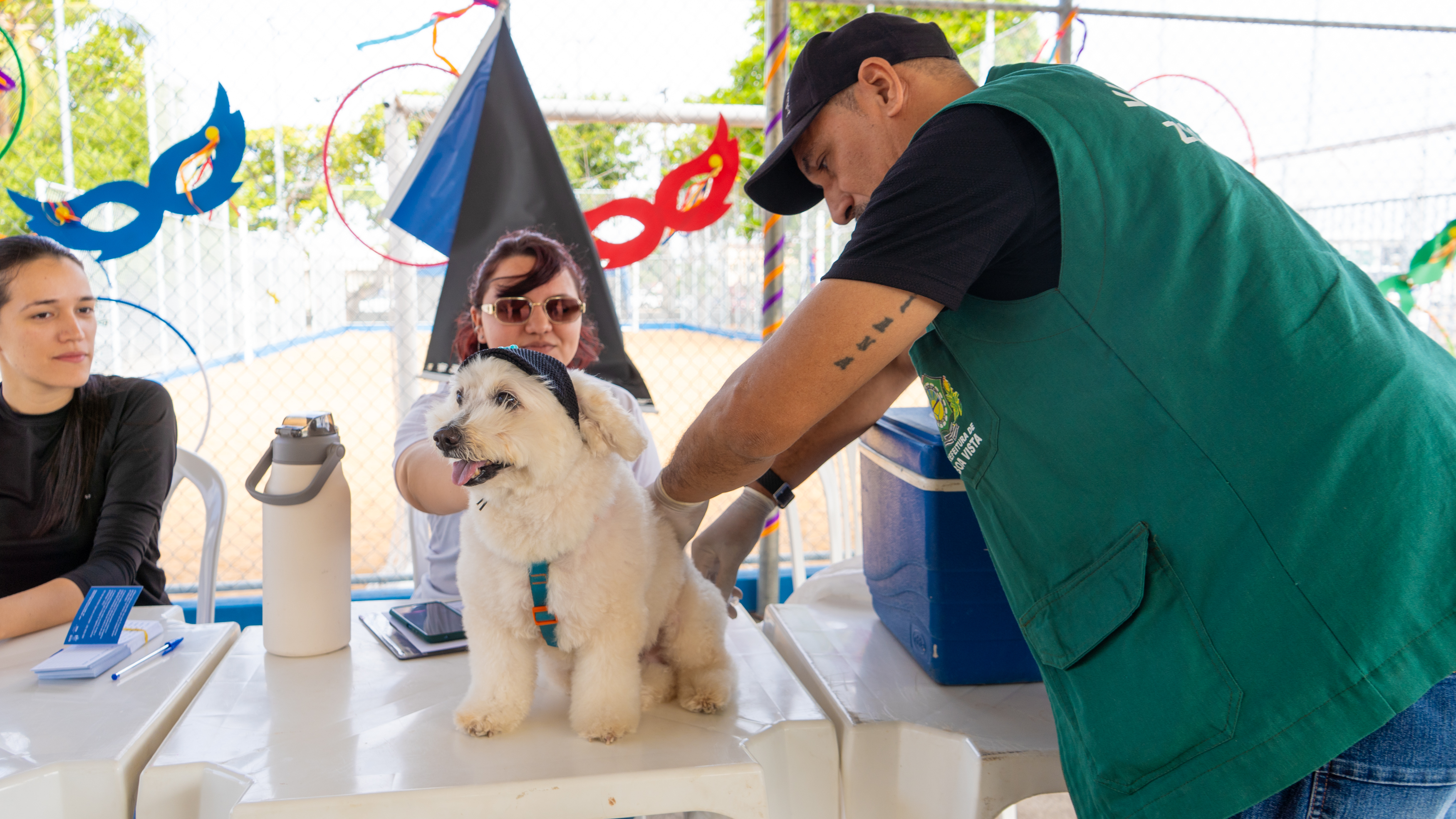 imagem colorida em ambiente externo de um homem vacinando um cachorro branco de pequeno porte encima de uma mesa branca.
