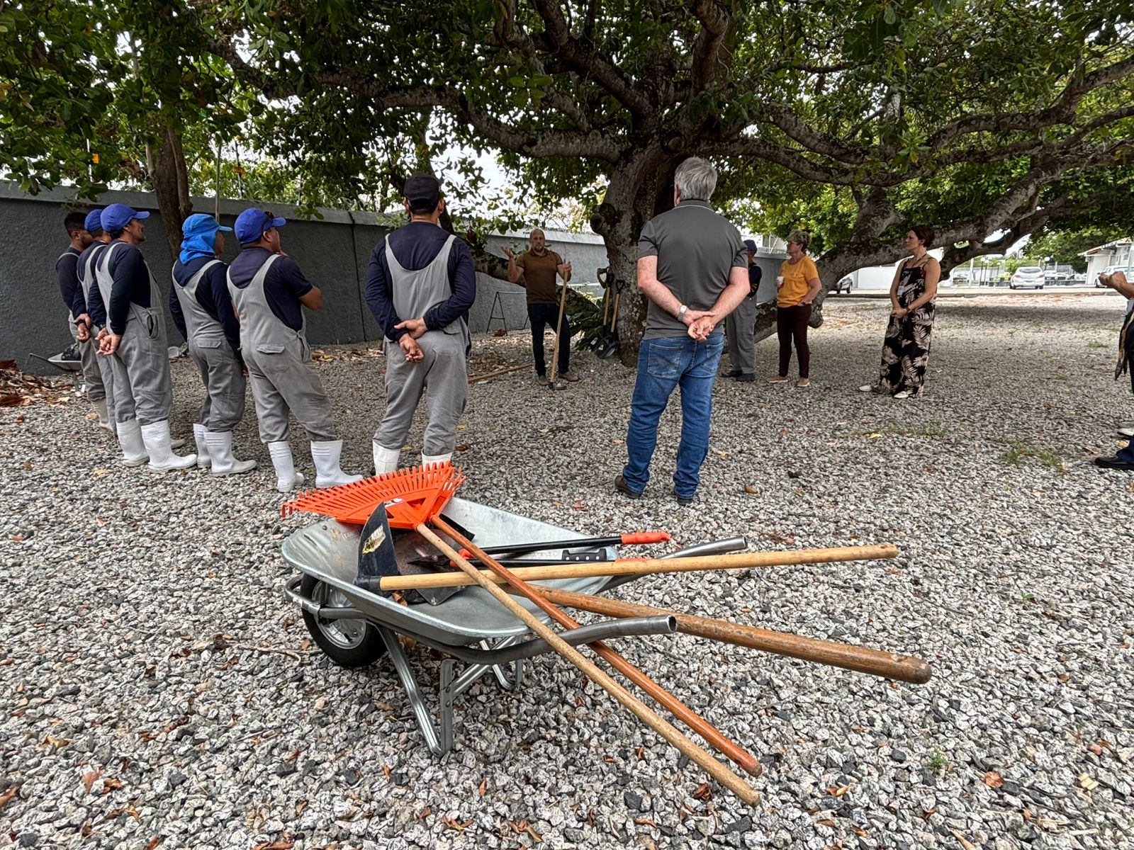 Dois homens com macacão cinza e botas brancas inclinam um carrinho de mão, despejando terra escura e úmida sobre uma pilha de folhas secas. Ao fundo, outros trabalhadores observam a demonstração prática de compostagem ao ar livre. Dois homens com macacão cinza e botas brancas inclinam um carrinho de mão, despejando terra escura e úmida sobre uma pilha de folhas secas. Ao fundo, outros trabalhadores observam a demonstração prática de compostagem ao ar livre.