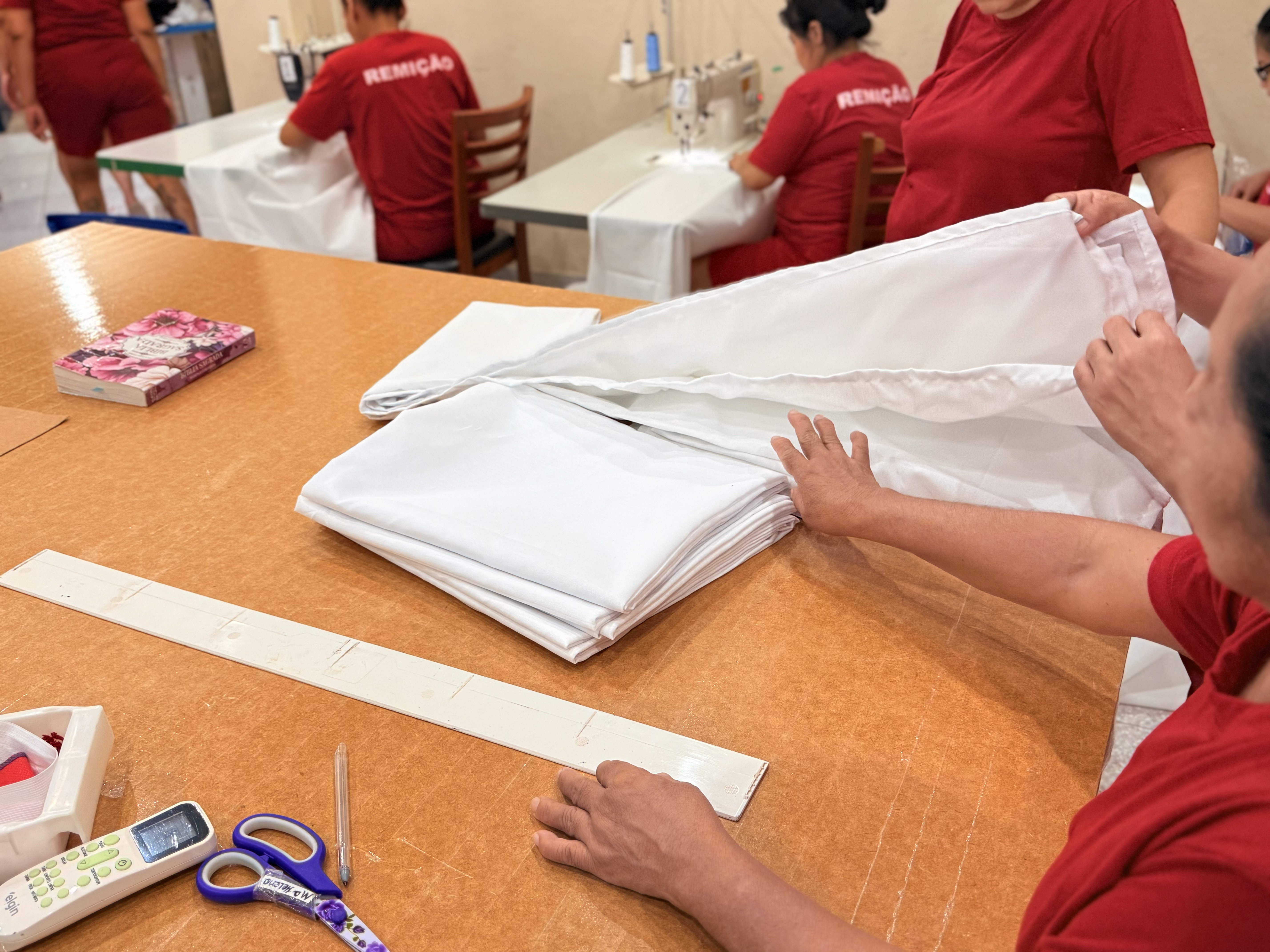   Imagem colorida mostra seis mulheres desfocadas em um ateliê emfileiradas em mesas coom maquinas de costura.