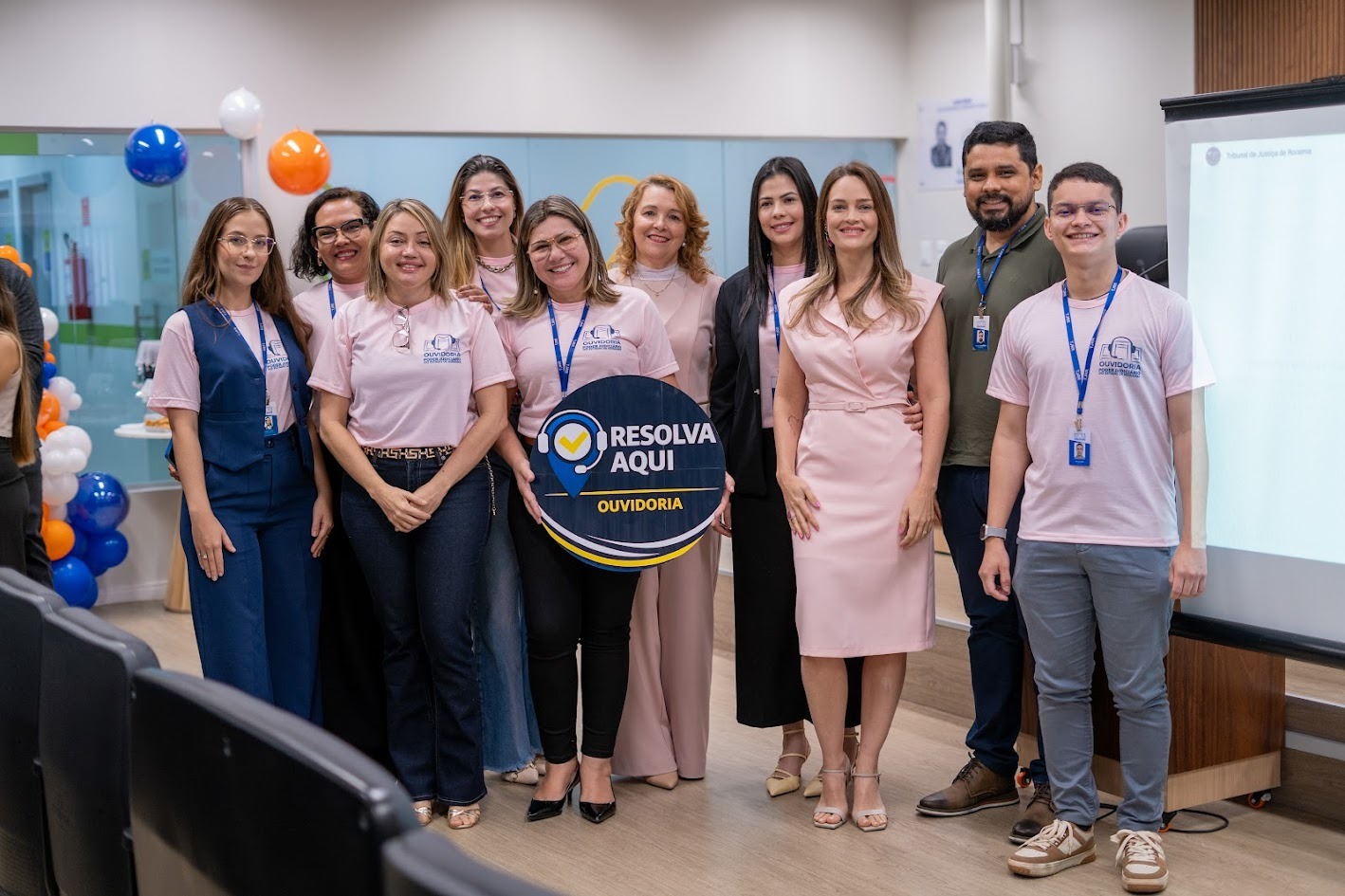  imagem colorida de um grupo de pessoas em um auditório, posando para foto. Parte do grupo veste camiseta rosa com crachá. Ao centro, uma pessoa segura uma placa azul com o texto “Resolva Aqui – Ouvidoria”. Ao fundo, há balões nas cores azul, laranja e branco e um telão de projeção.