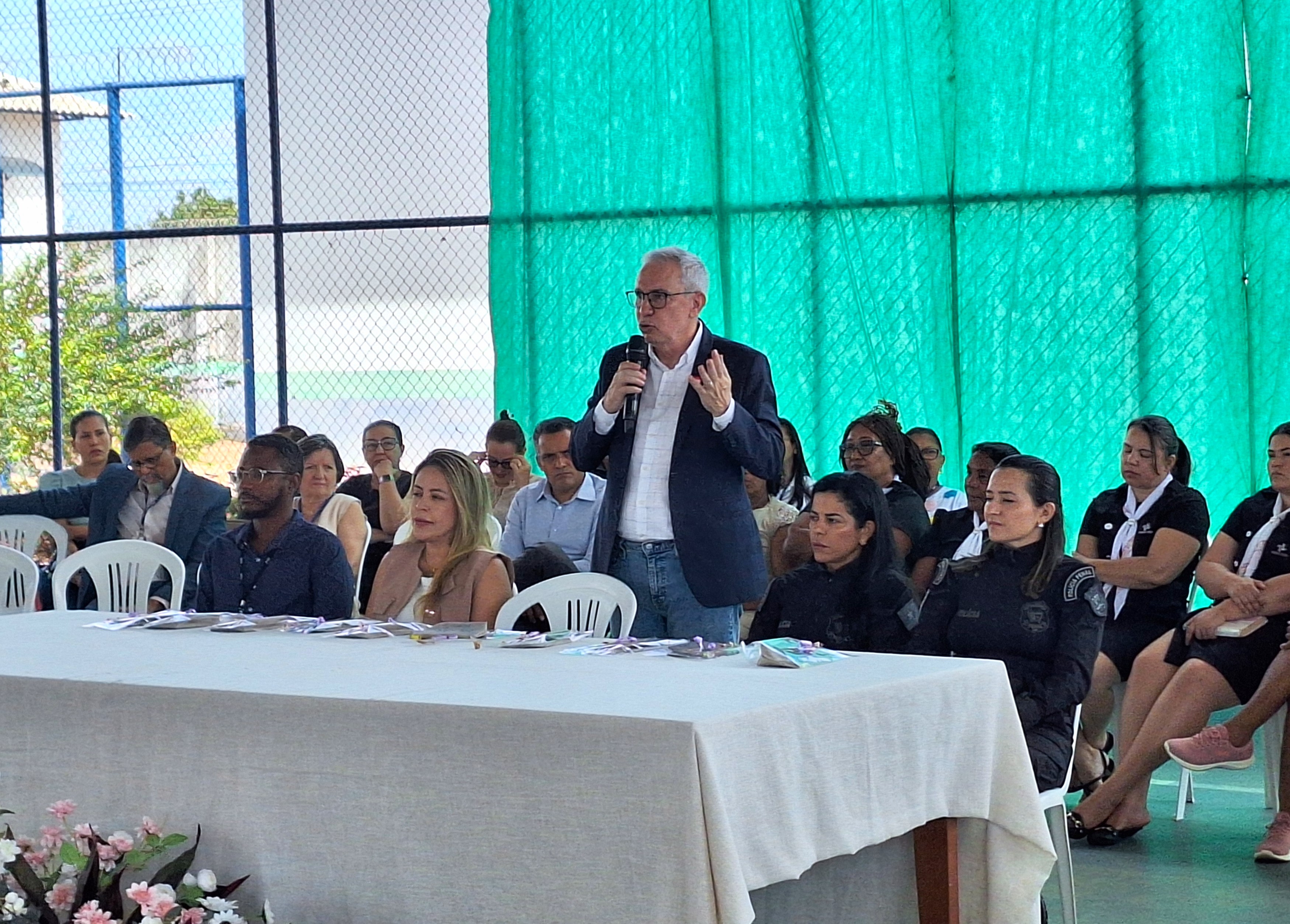 Foto colorida mostra o Desembargador Almiro Padilha fala ao microfone enquanto está em pé próximo a uma mesa. Ao redor, várias pessoas estão sentadas assistindo ao pronunciamento, incluindo mulheres com uniforme escuro.